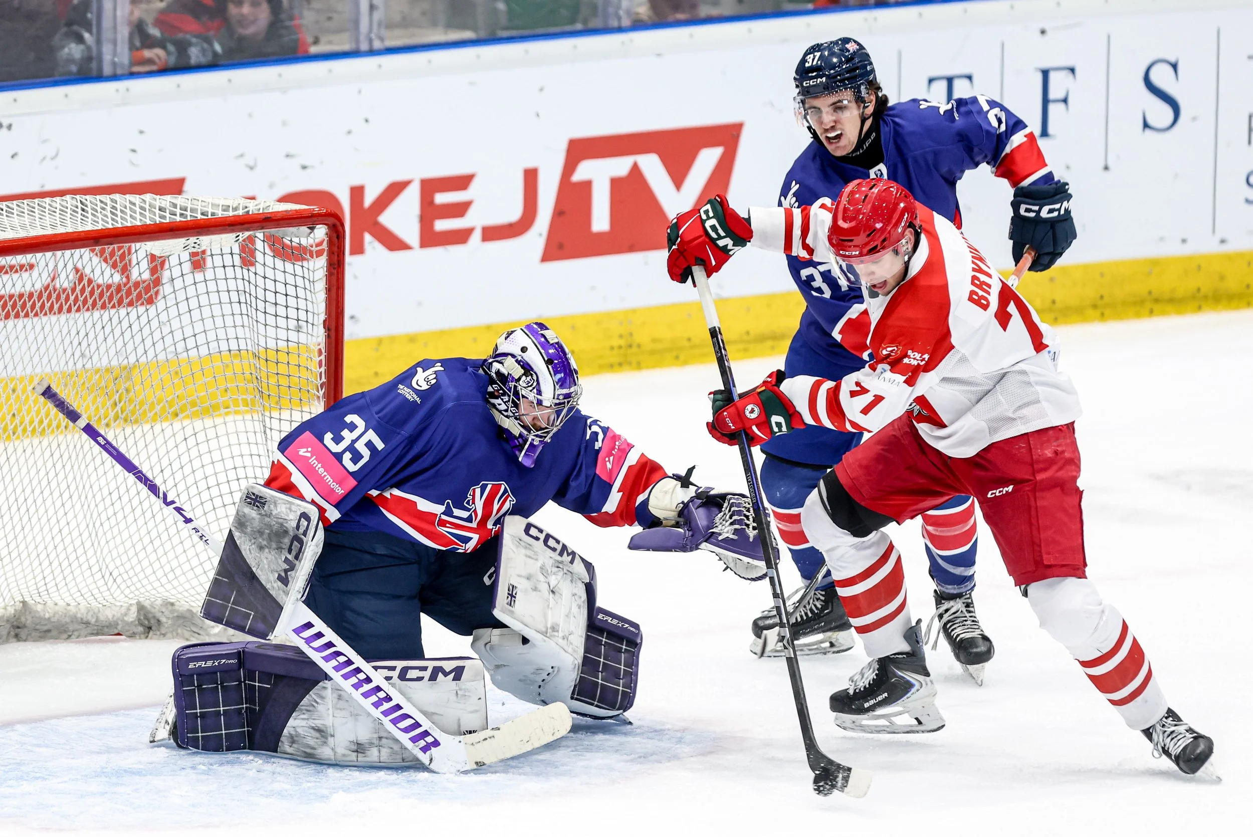 Hockey players competing for the puck in front of the goal, with the goalie crouched and prepared to block the shot.