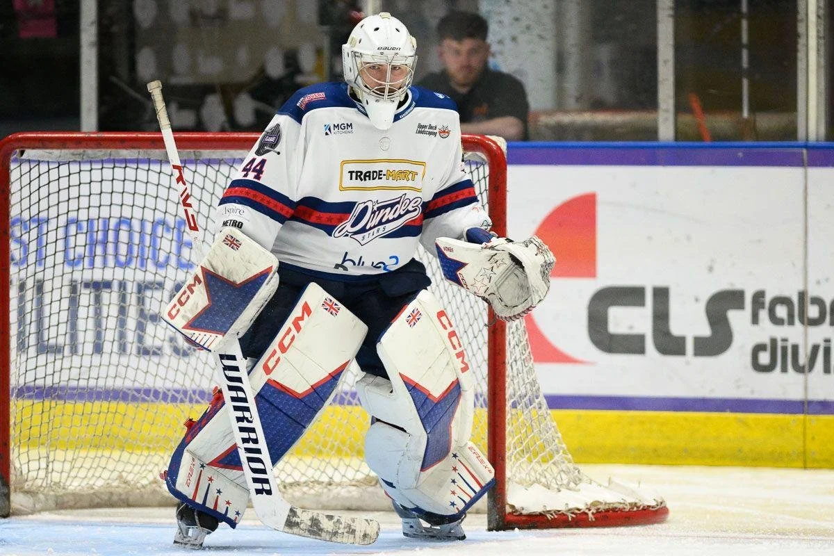 An ice hockey goalie in full gear stands in front of the net on the ice rink during a game. The goalie is wearing a white, blue, and red uniform with the number 44, and is holding a blocker in one hand and a glove in the other.