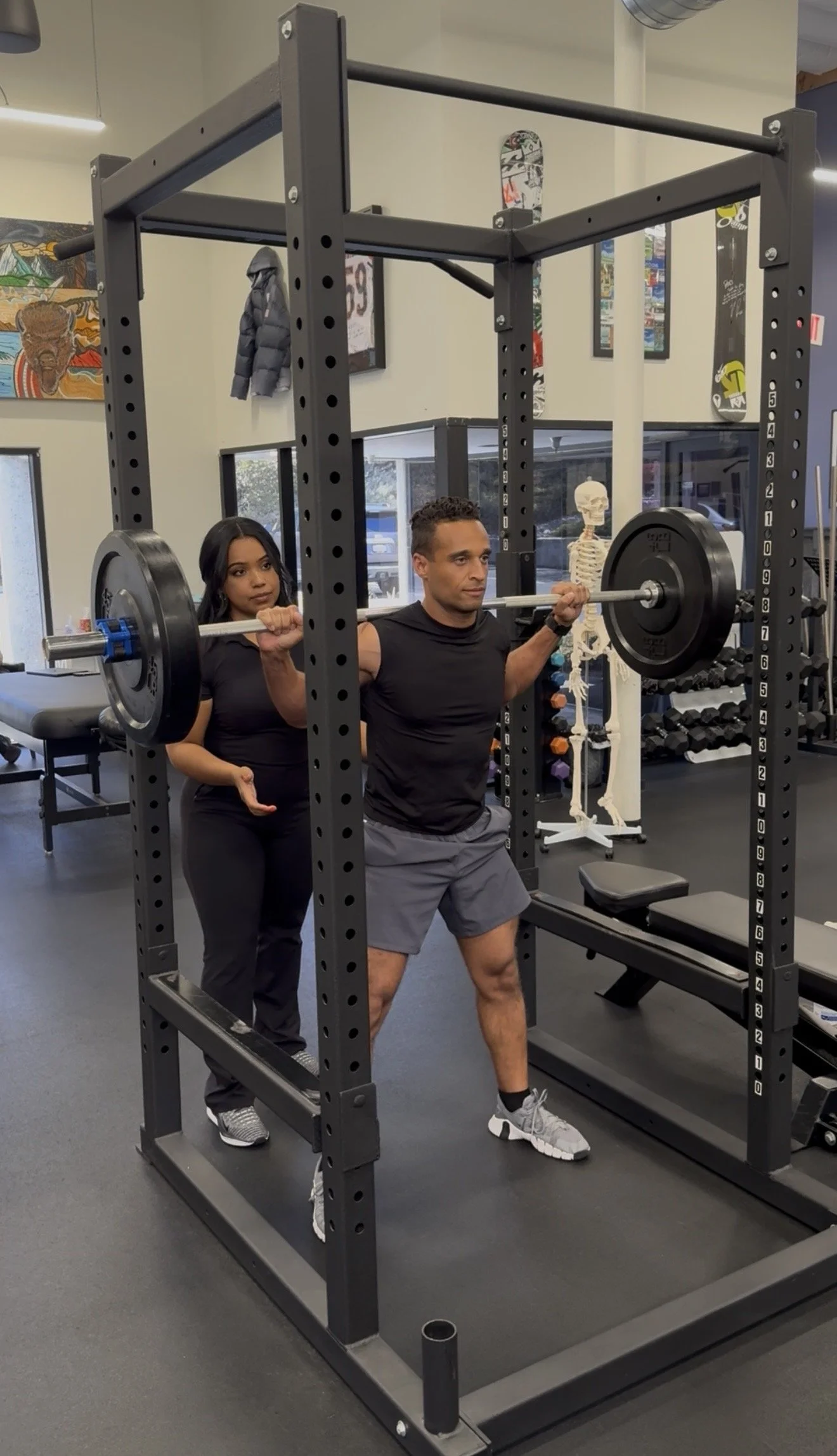 A man is performing a squat exercise with a barbell while a woman stands behind him spotting. They are in a gym with various fitness equipment, a skeleton model, skateboard wall art, and large windows.