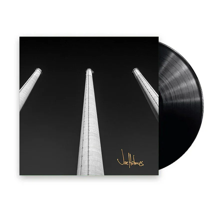 Black and white photo of three tall industrial smokestacks reaching into a dark sky, with a vinyl record partially visible behind the album cover. The album has a signature that reads 'Joe Holmes' in gold.