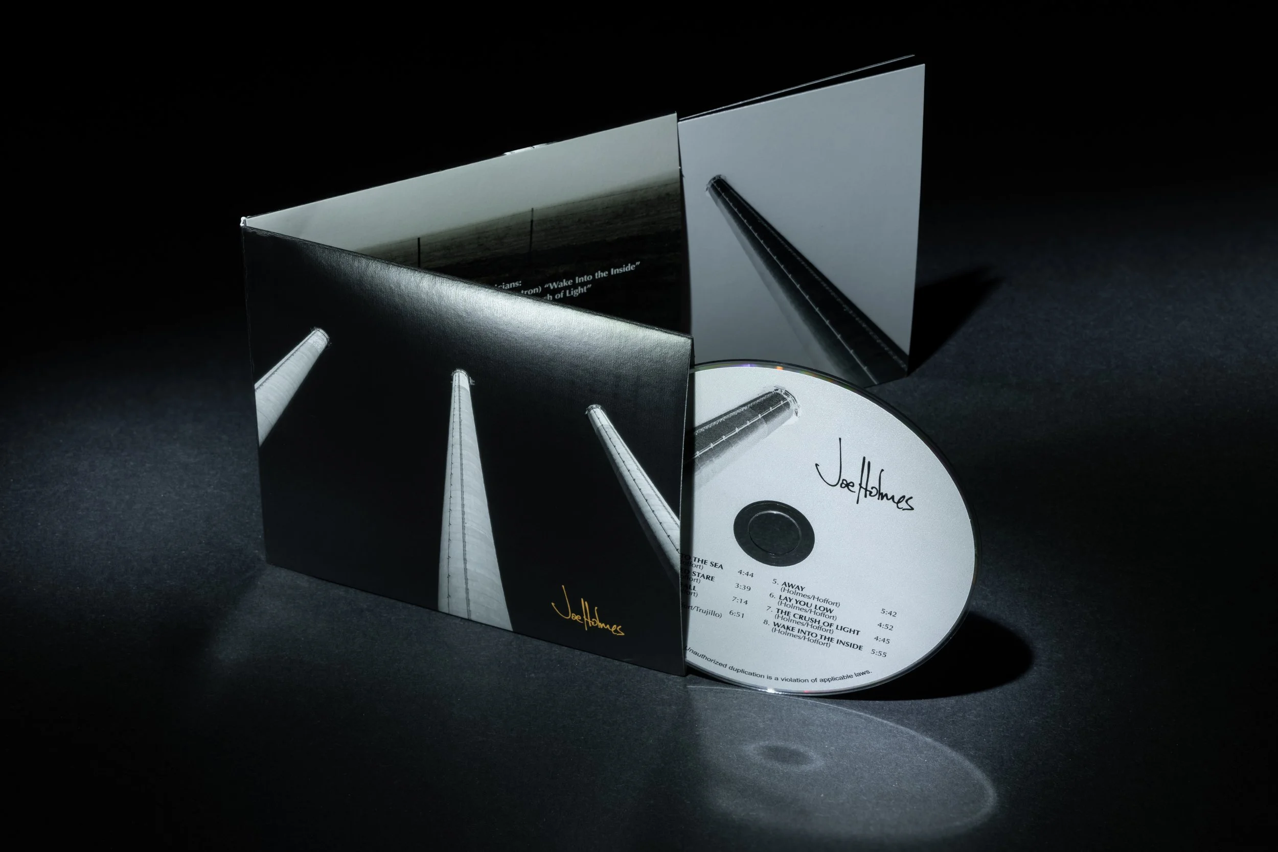 Open CD case with a CD inside and an accompanying booklet or sleeve, both featuring a black-and-white cover with tall, narrow columns. The CD has handwritten text that reads 'Joe Holmes' and song list.