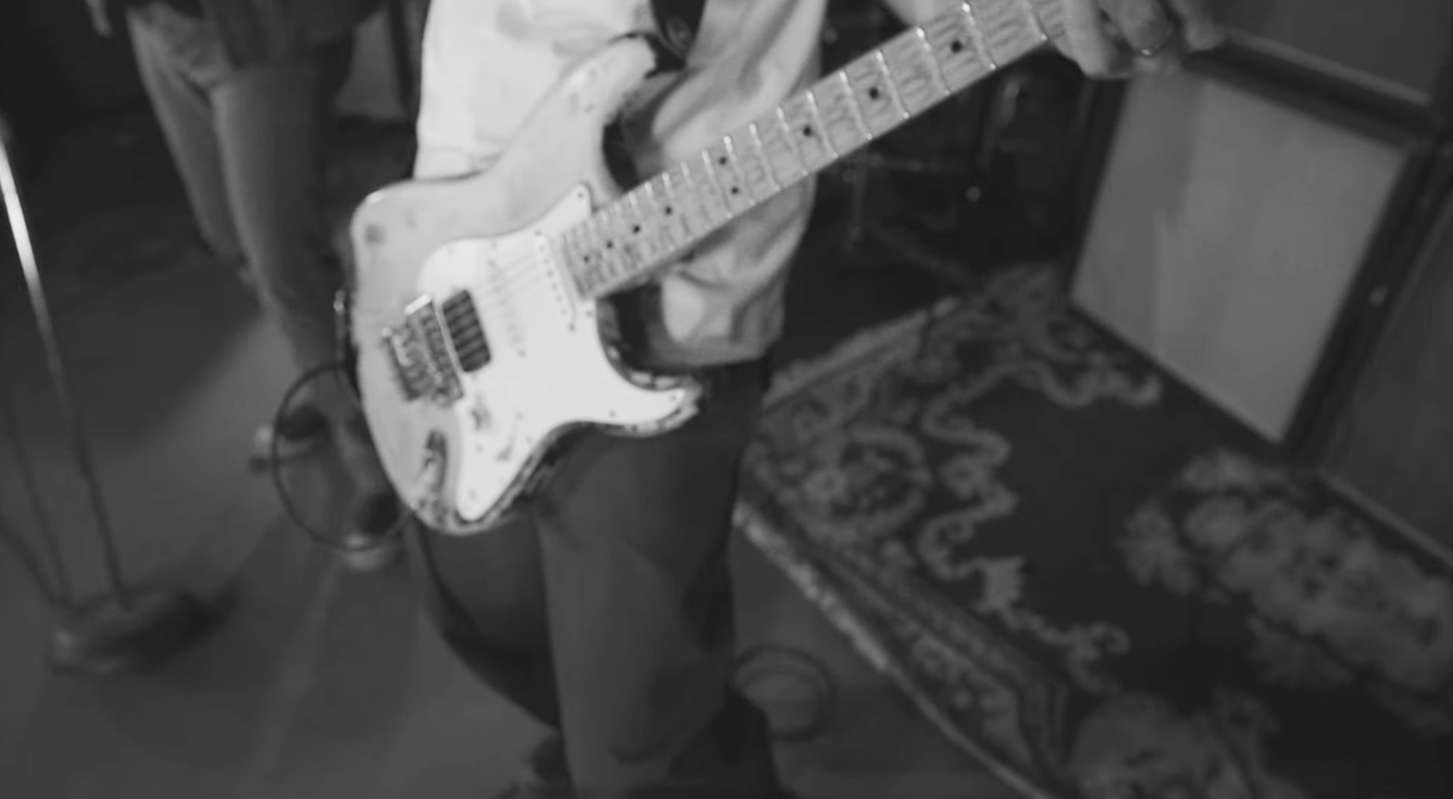 Black and white image of an electric guitar resting on a chair, with an amplifier and a patterned rug visible nearby.