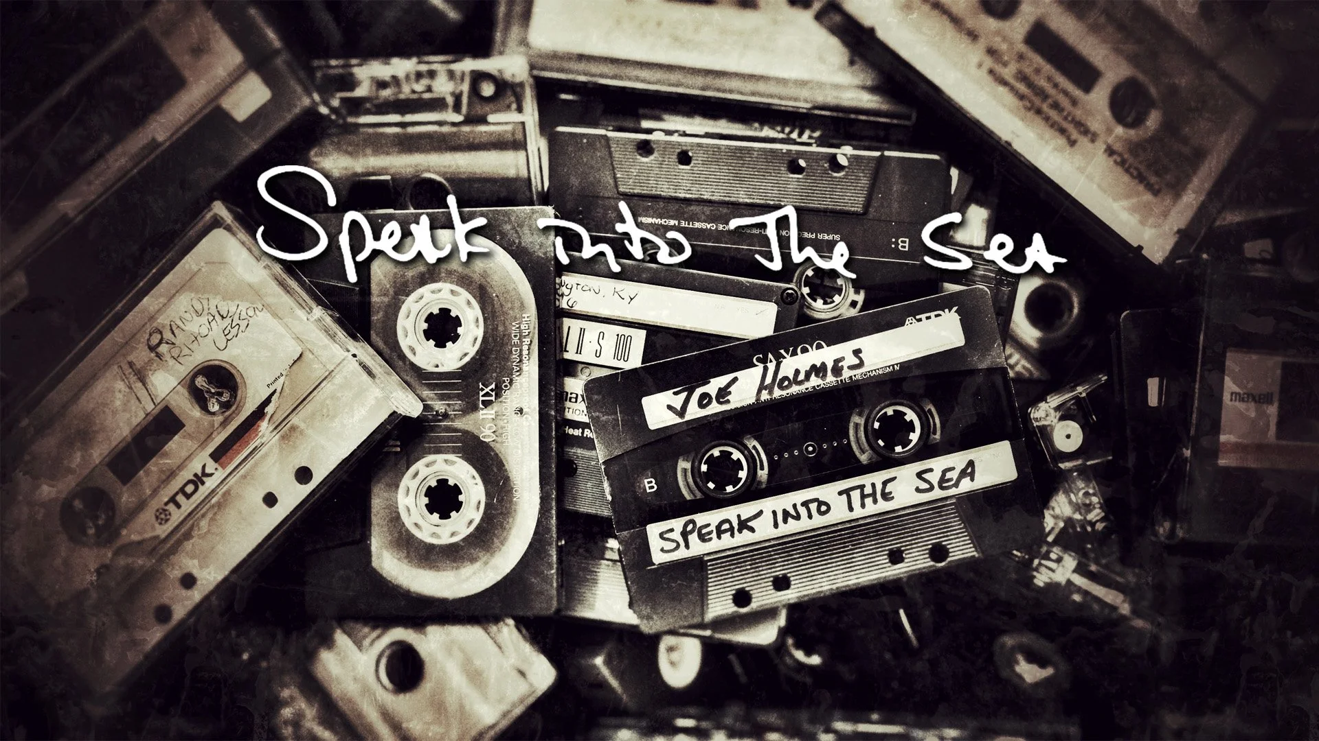 A collection of vintage cassette tapes, some with handwritten labels including "Joe Holmes" and "Speak Into the Sea," stacked on top of each other, with The words "Speak Into the Sea" written across the center of the image.