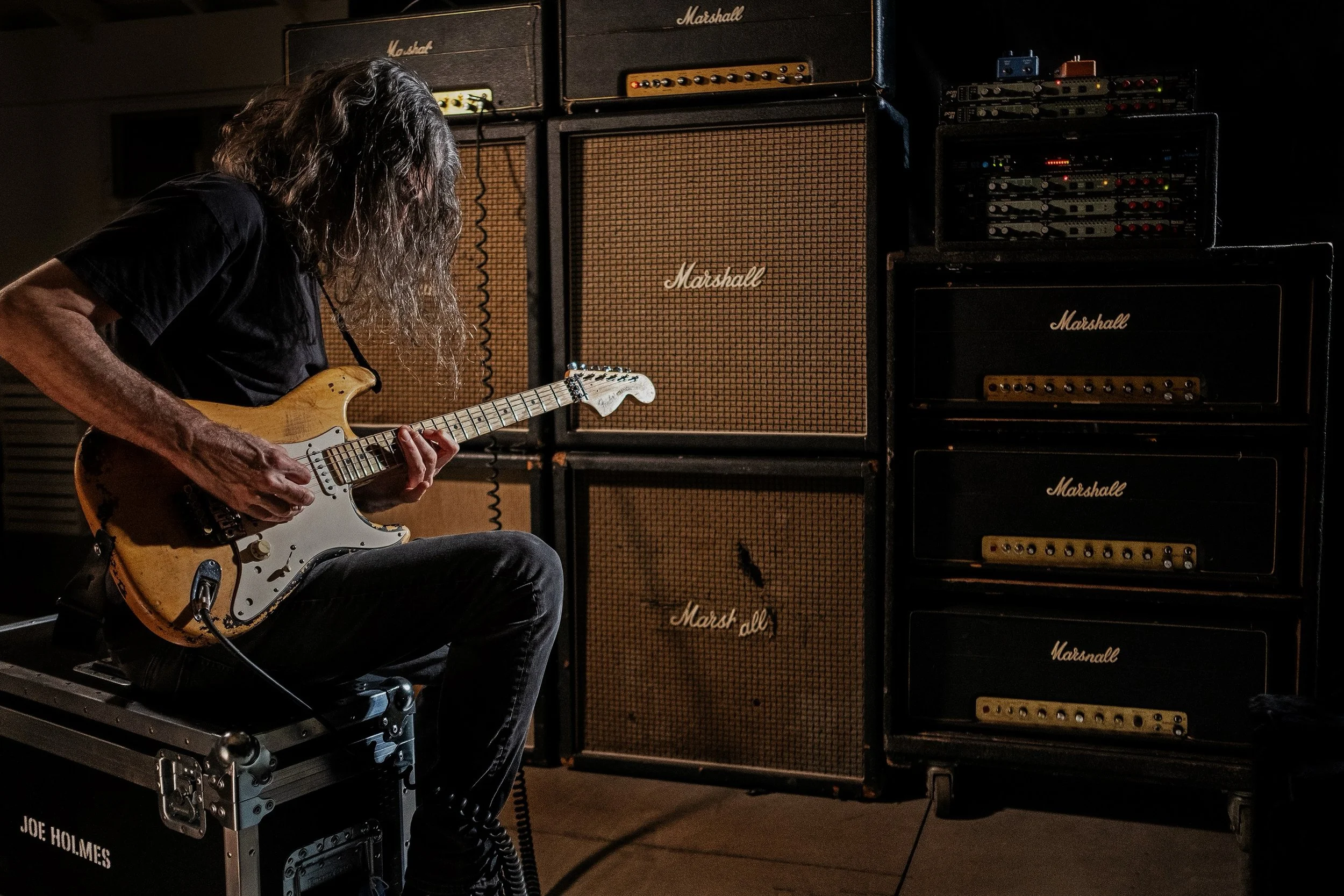 A person with long curly hair playing an electric guitar in front of stacks of Marshall amplifiers and audio equipment in a dark room.