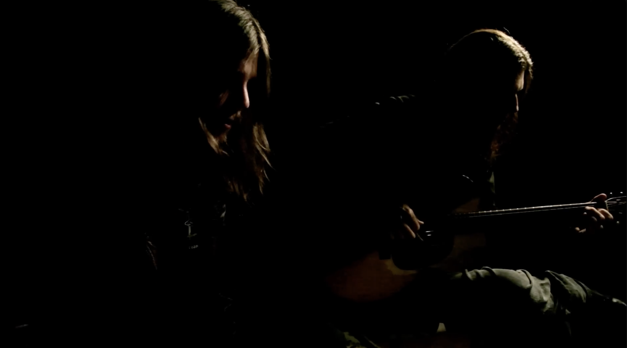 Two women sitting and playing guitars in dim lighting with their faces partially visible.