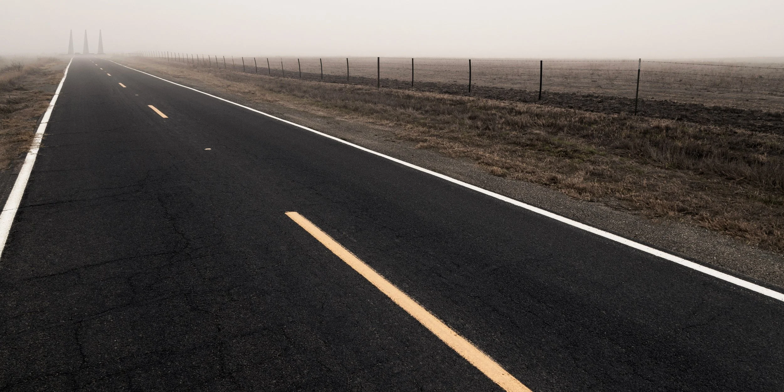 Empty black asphalt road stretching into foggy horizon, bordered by white lines and yellow dashed centerlines, with dry grassy fields and a barbed wire fence on the right side.