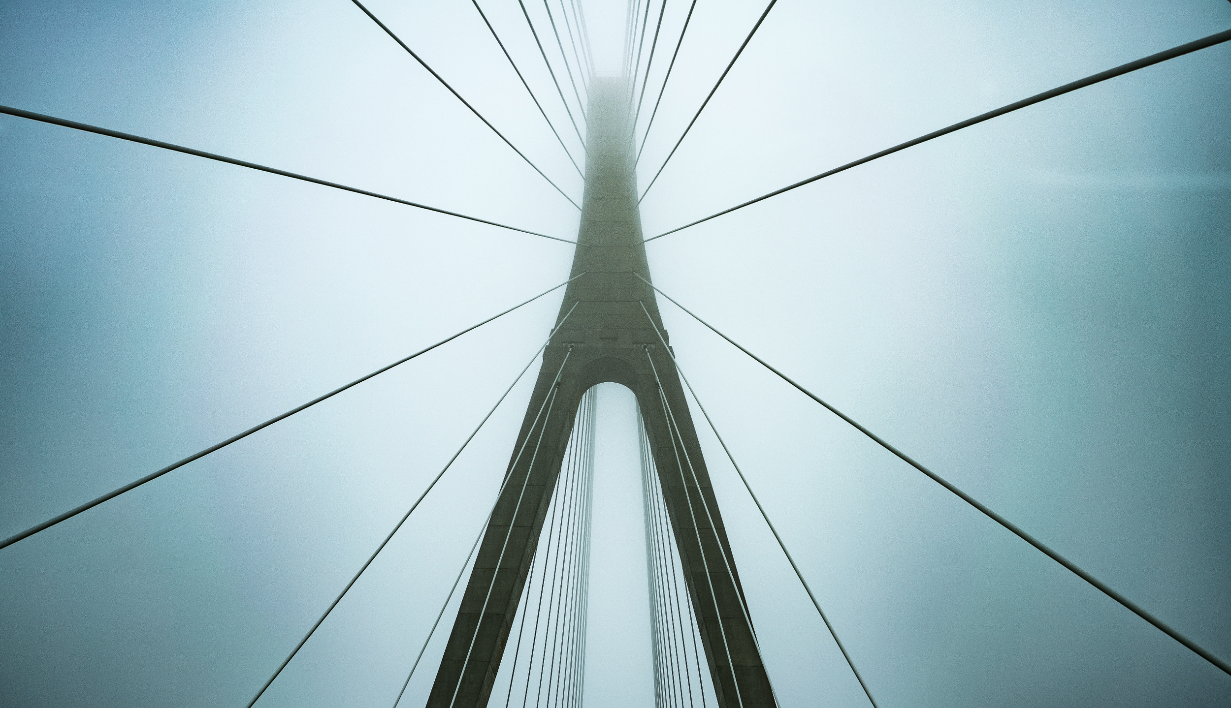 Upward view of a bridge tower with suspension cables in a foggy or overcast sky.