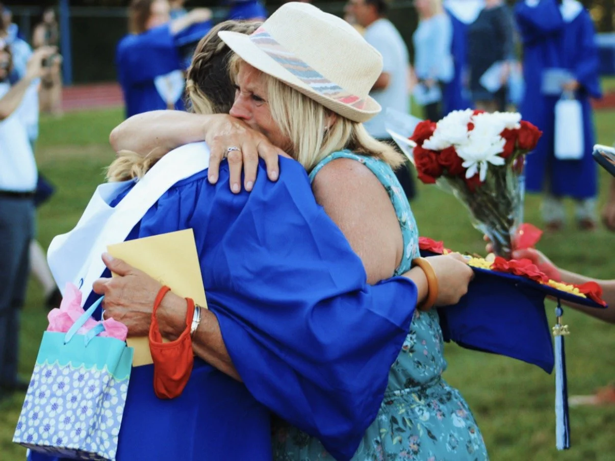 A woman in a floral dress and hat hugs a graduate wearing a blue cap and gown, holding a bouquet of red and white flowers and a yellow envelope, at an outdoor graduation ceremony.