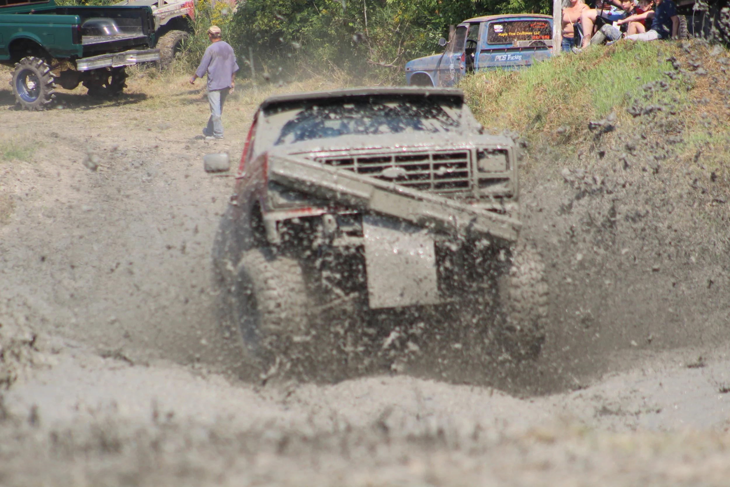 A muddy off-road vehicle driving through a dirt trail at a rugged outdoor event with spectators and other vehicles in the background.
