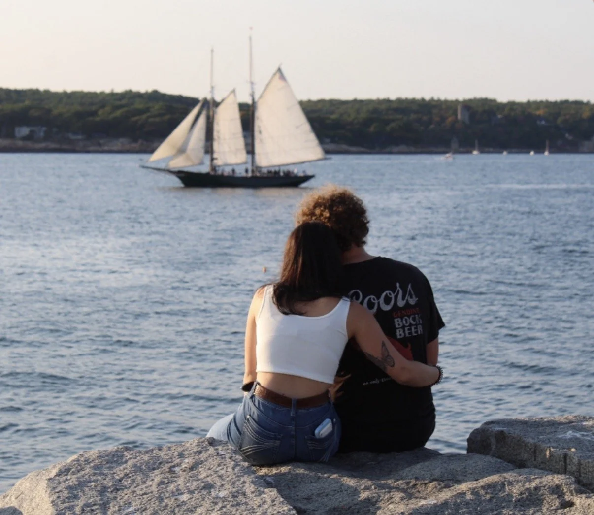 Couple sitting on rocks by the water, watching a sailing ship and smaller boats on the water with a distant shoreline and trees.