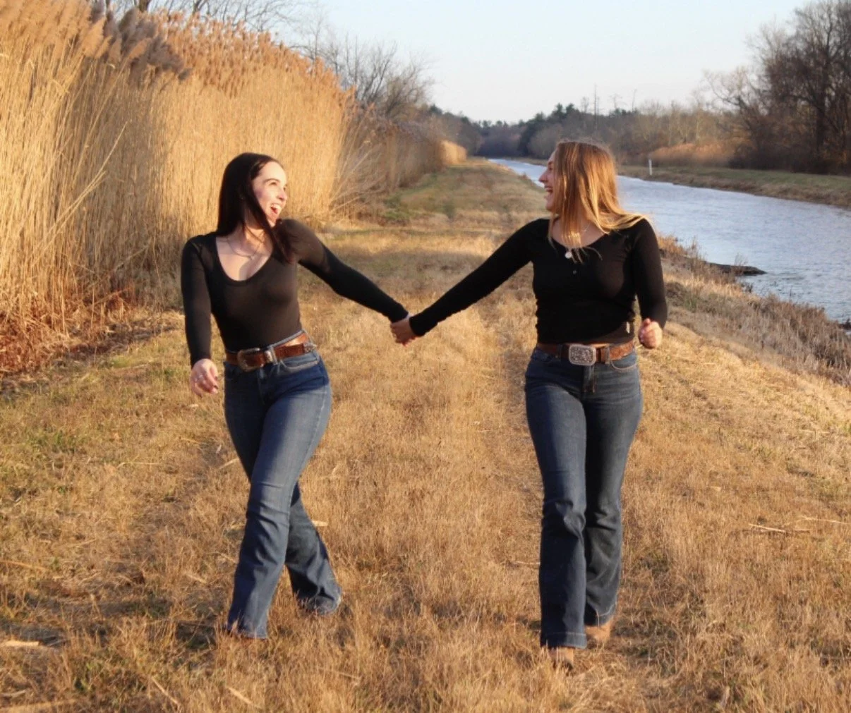 Two women in black shirts and jeans holding hands and smiling while walking along a grassy dirt path beside a river and tall dried reeds.