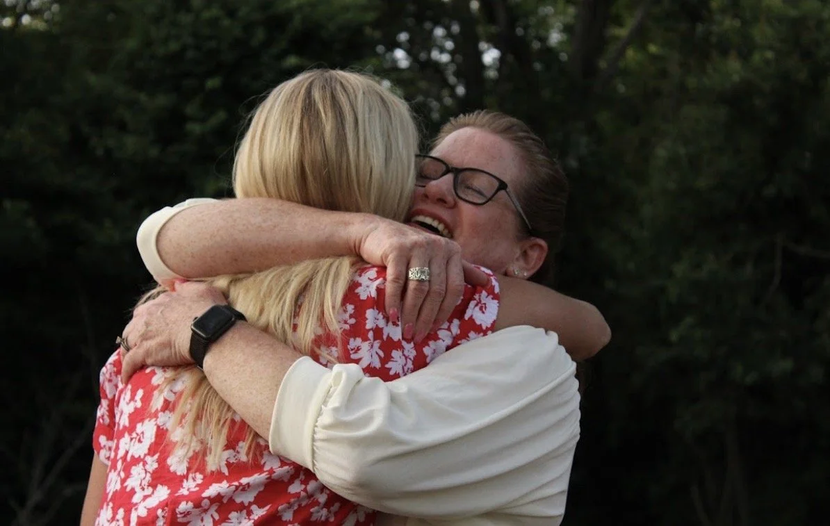 Two women hugging and one woman smiling with eyes closed.