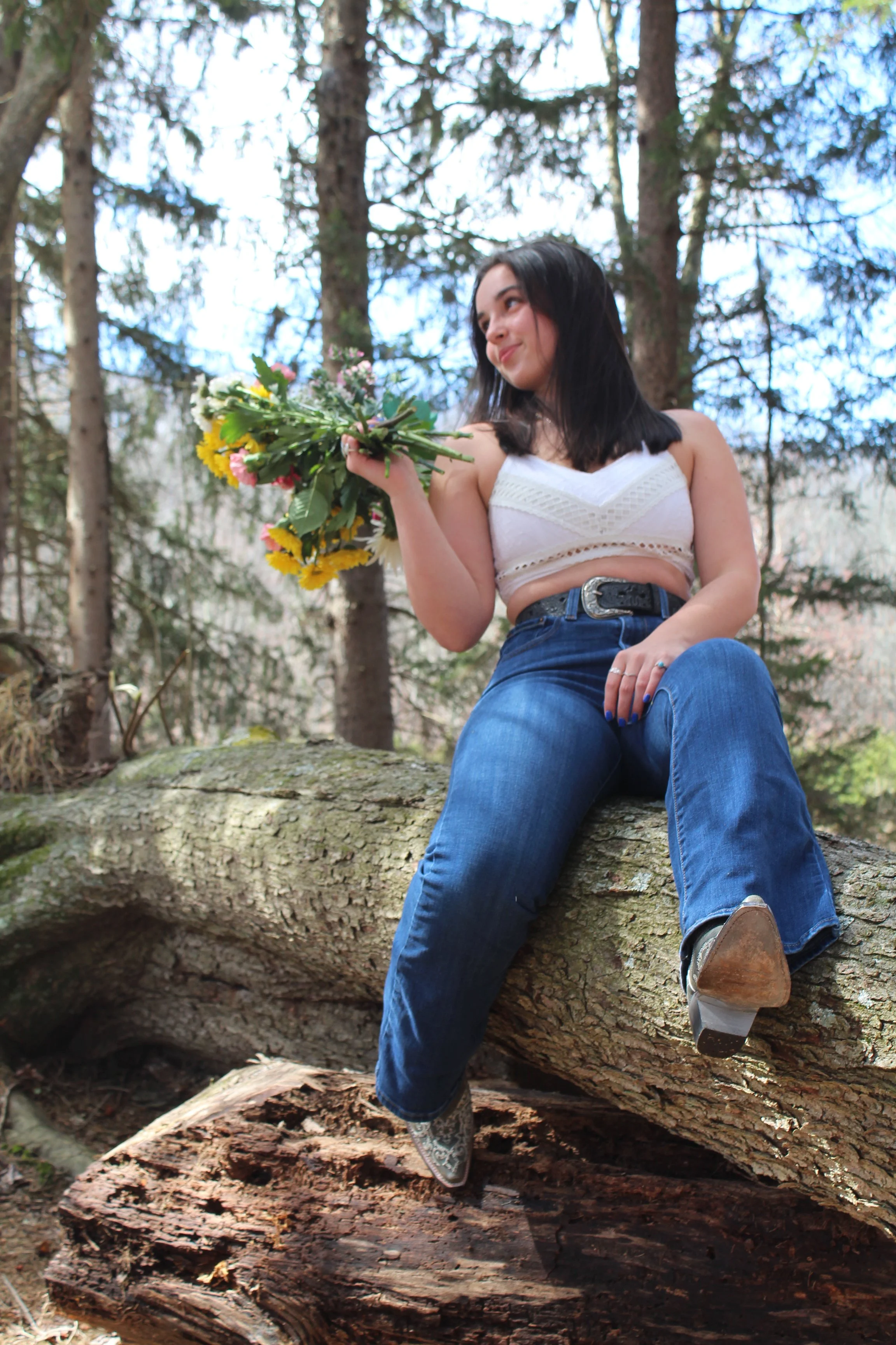 Young woman sitting on a fallen tree trunk in a forest, holding a bouquet of colorful flowers, smiling, wearing a white crop top and jeans.