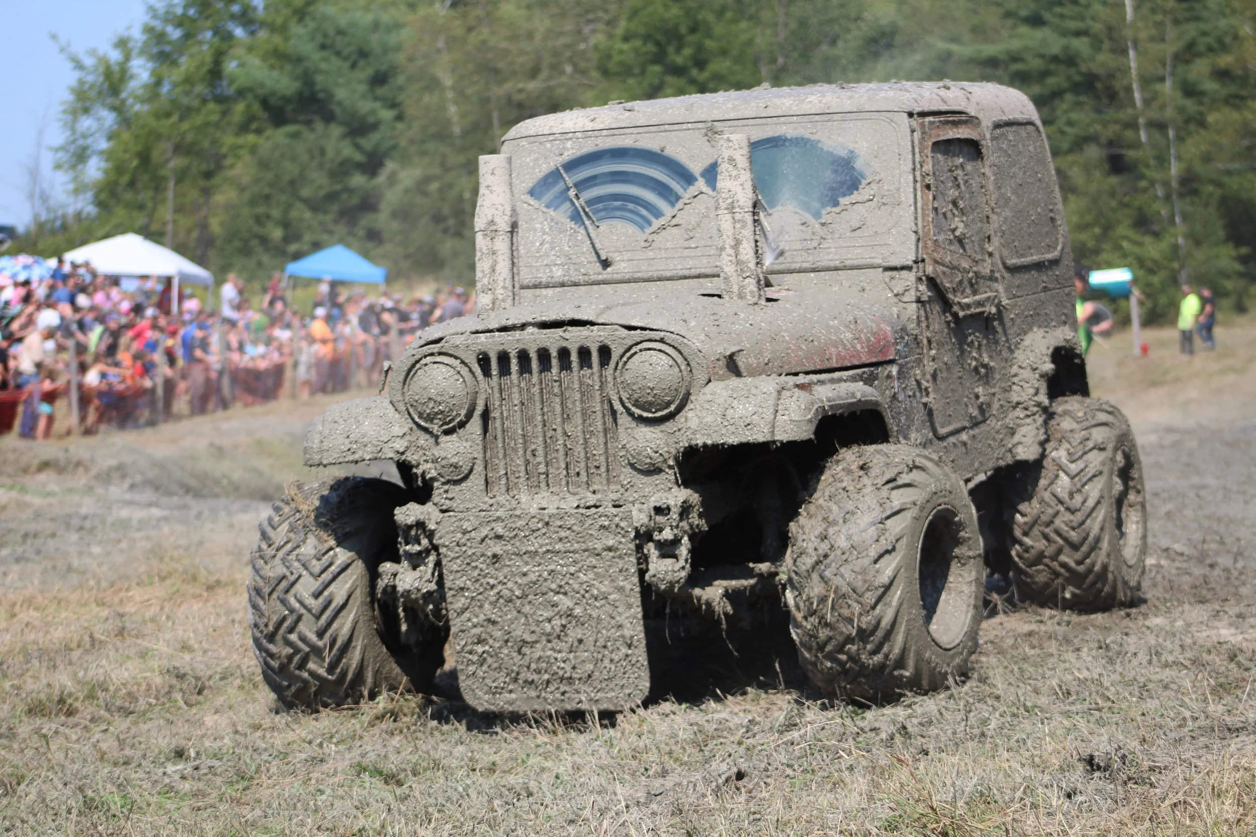 A muddy, off-road vehicle with shattered windshield, muddy tires, and a dusty exterior, on a grassy field during an outdoor event with people and tents in the background.