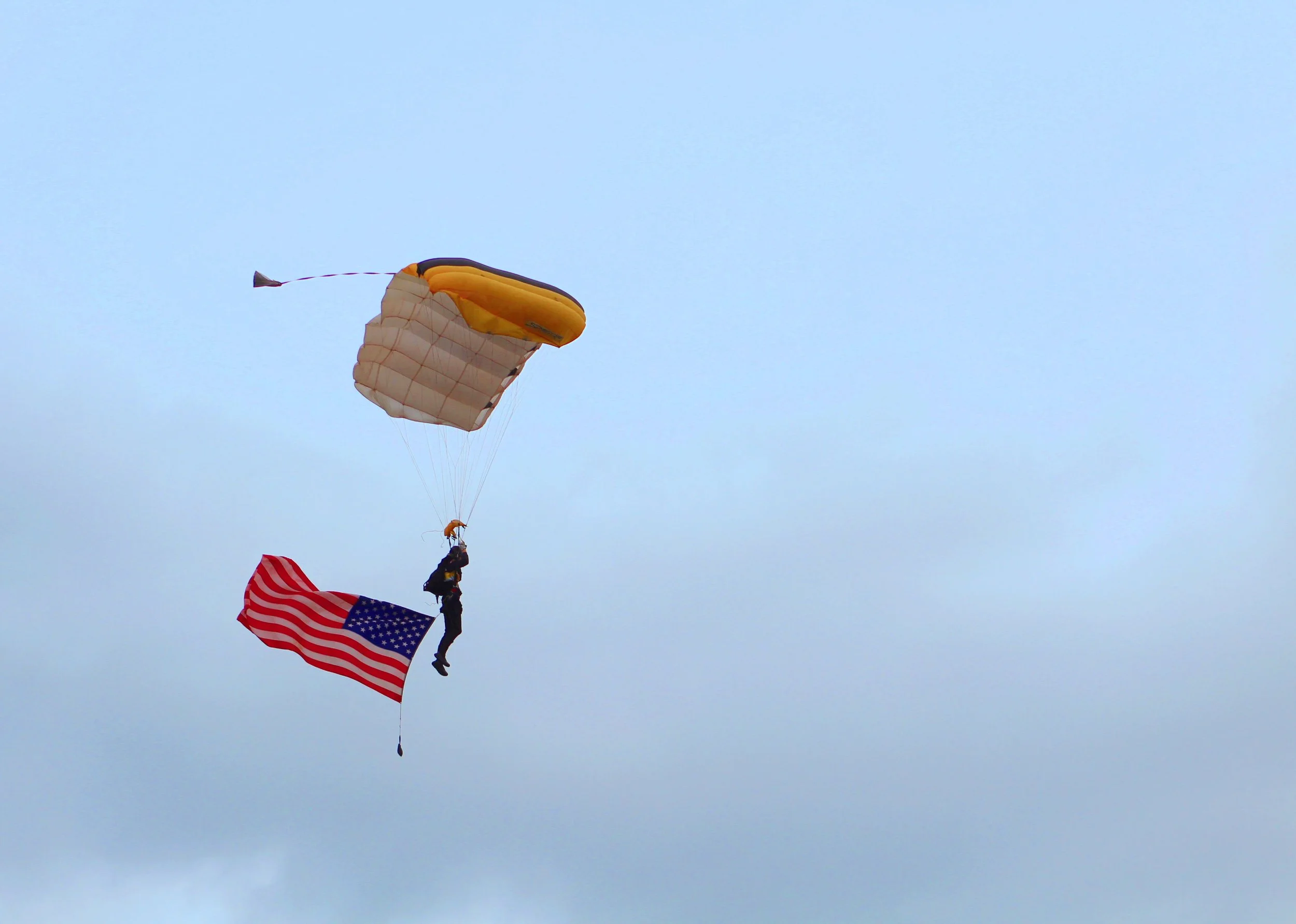 A person hanging from a parachute that has an American flag attached to the bottom, flying in the sky on a sunny day.