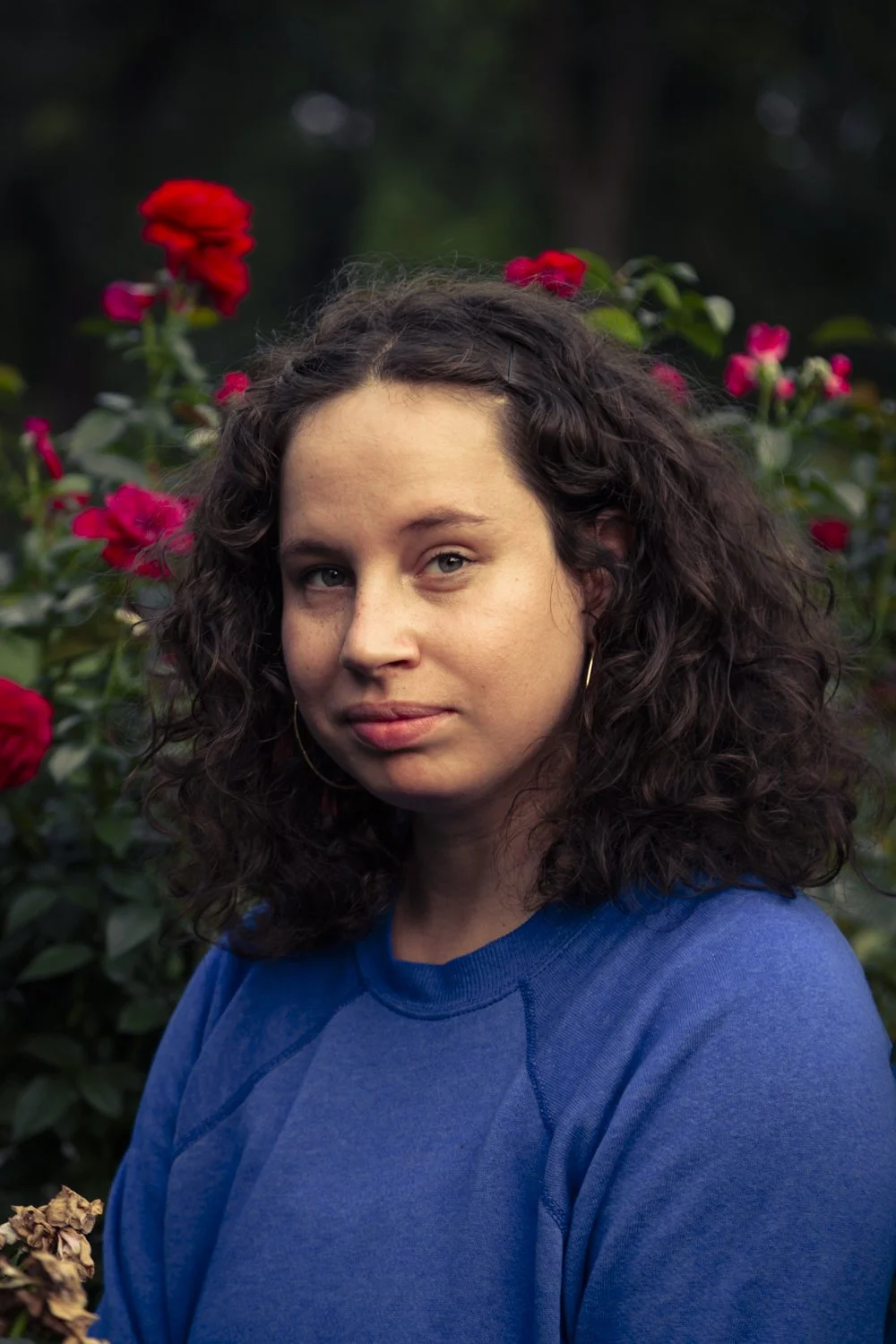 A young woman with curly dark hair and hoop earrings standing outdoors in front of pink and red flowers, wearing a blue shirt.
