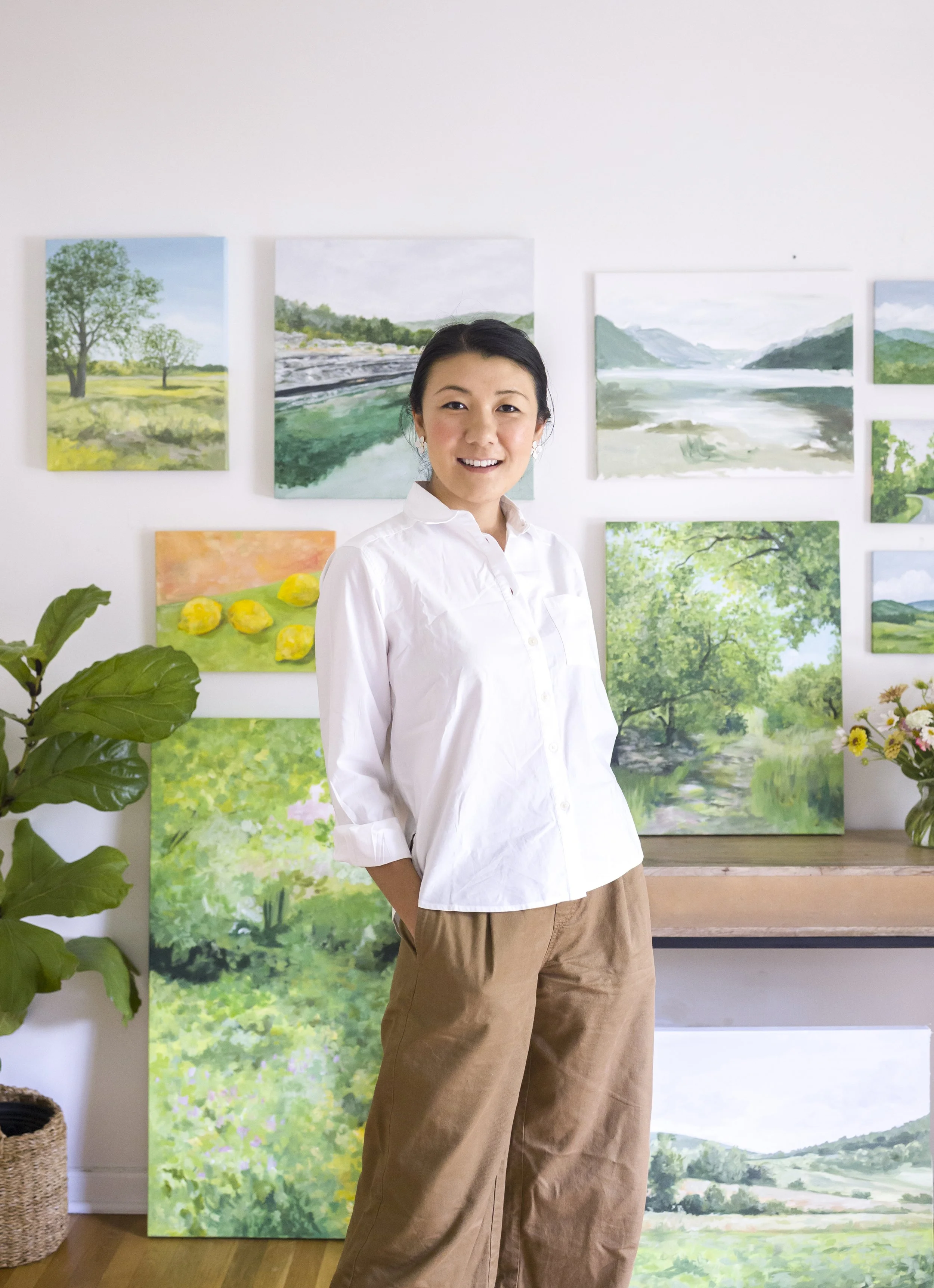 A woman in a white shirt and brown pants standing in an art gallery with landscape paintings on the wall behind her, and a plant on the left.