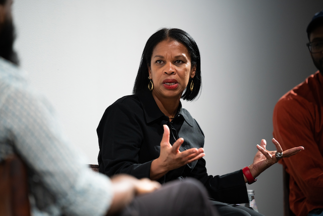 Woman with black hair and gold hoop earrings speaking during a discussion, gesturing with her hands, seated next to two other people in a panel or meeting setting.