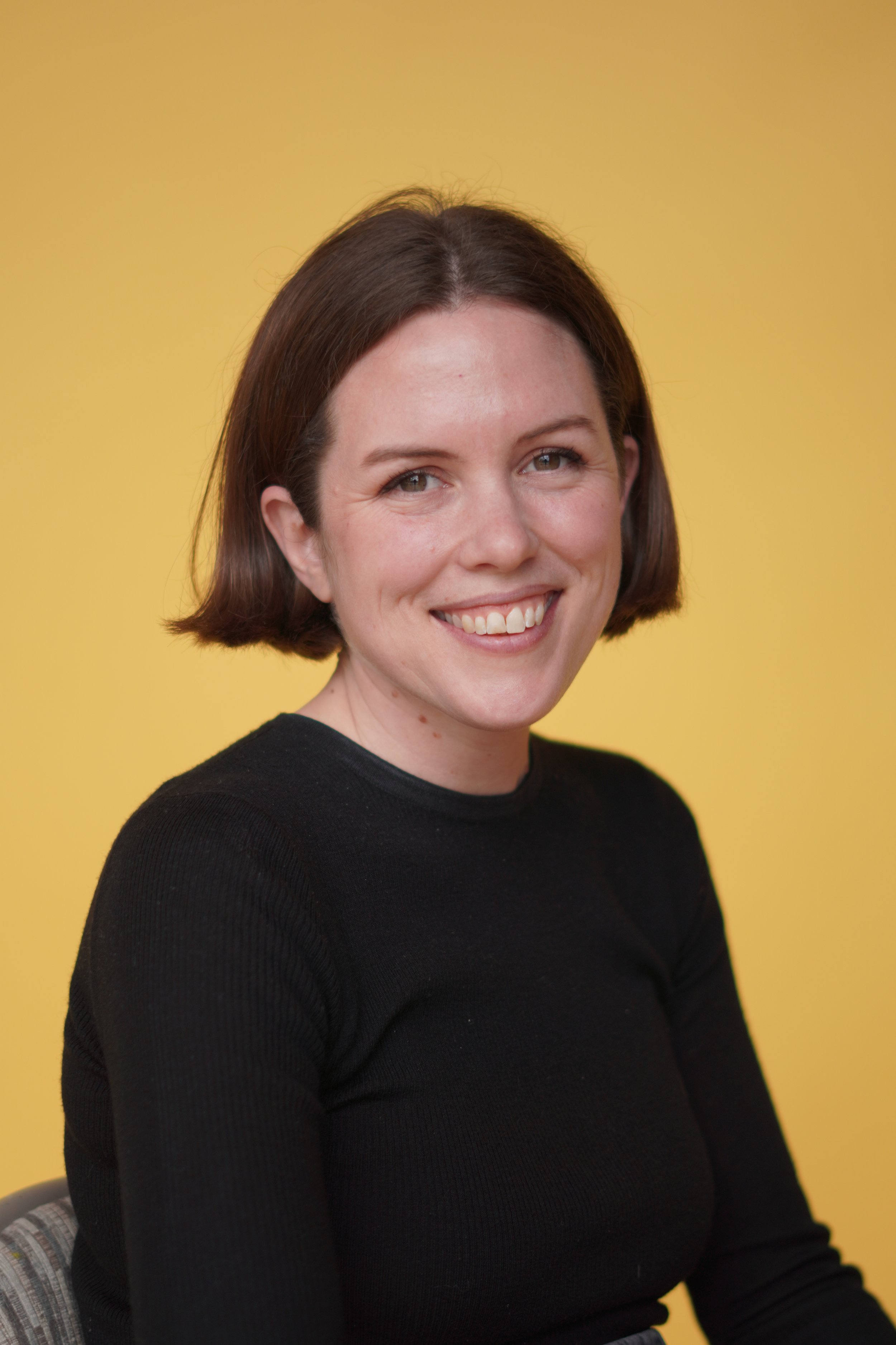 A woman with short brown hair smiling, wearing a black sweater, against a yellow background.