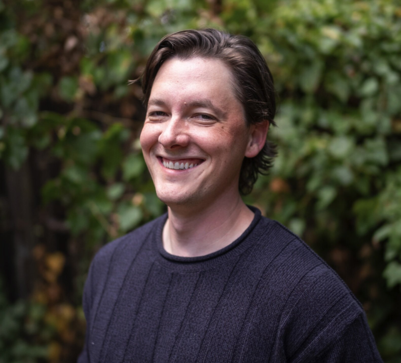 A smiling young man with dark hair and light skin, wearing a dark sweater, standing in front of a blurred green leafy background.