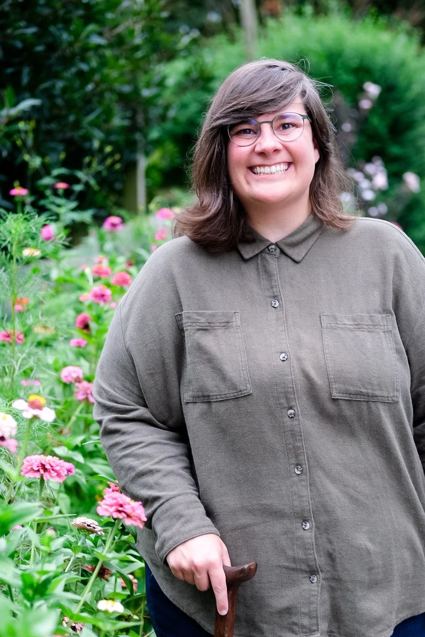 A smiling woman with glasses standing outdoors in a garden with pink flowers, holding a walking stick, wearing a green button-up shirt.