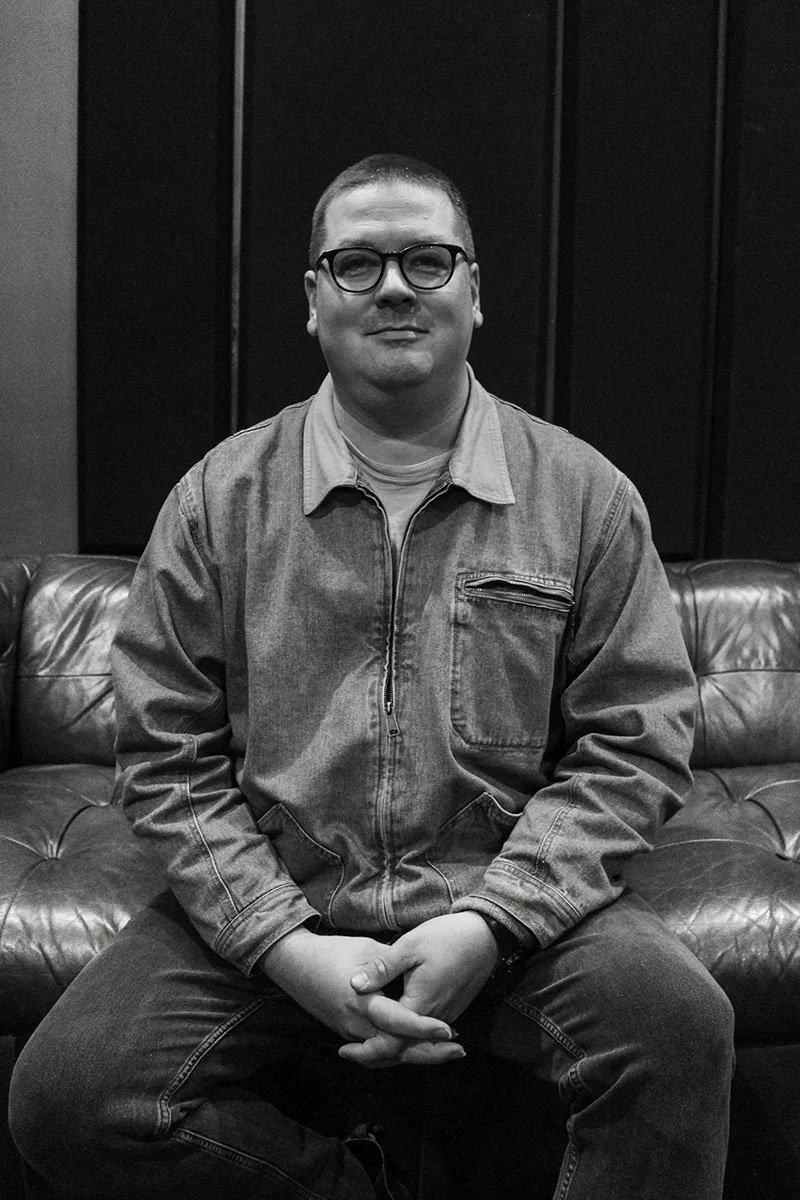 A man wearing glasses, a light-colored shirt, and a denim jacket, sitting on a leather couch in a dimly lit room with dark walls.