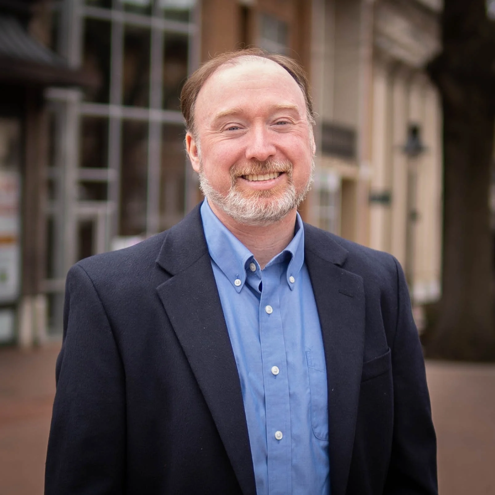 A man with a beard and mustache, wearing a dark blazer and a light blue button-up shirt. He is smiling and standing outdoors with wooden structures and windows in the background.