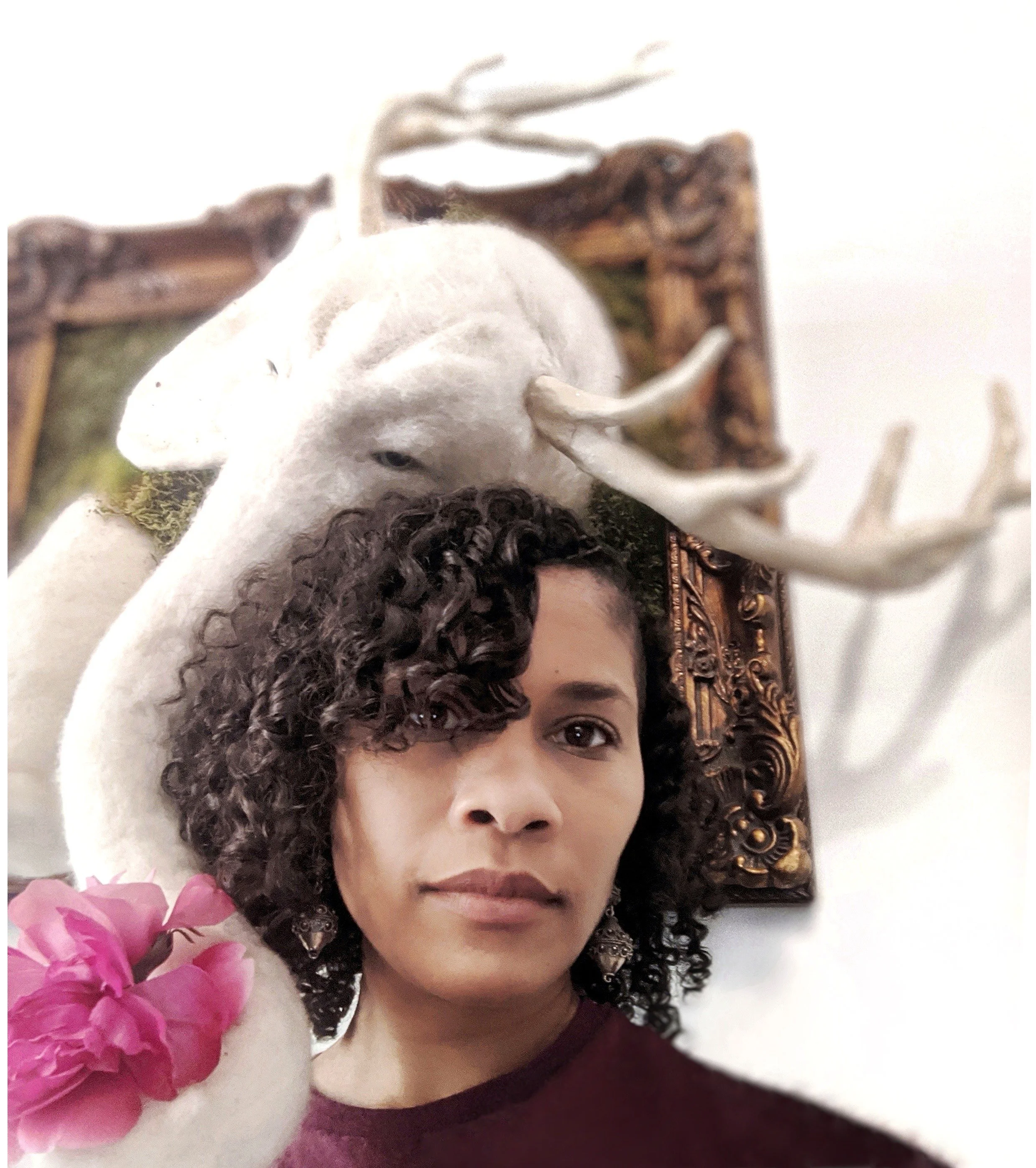 A close-up portrait of a woman with curly hair, wearing earrings and a maroon top, lying down with a smile. Behind her, there is a framed artwork, and in the foreground, a bunch of pink flowers.