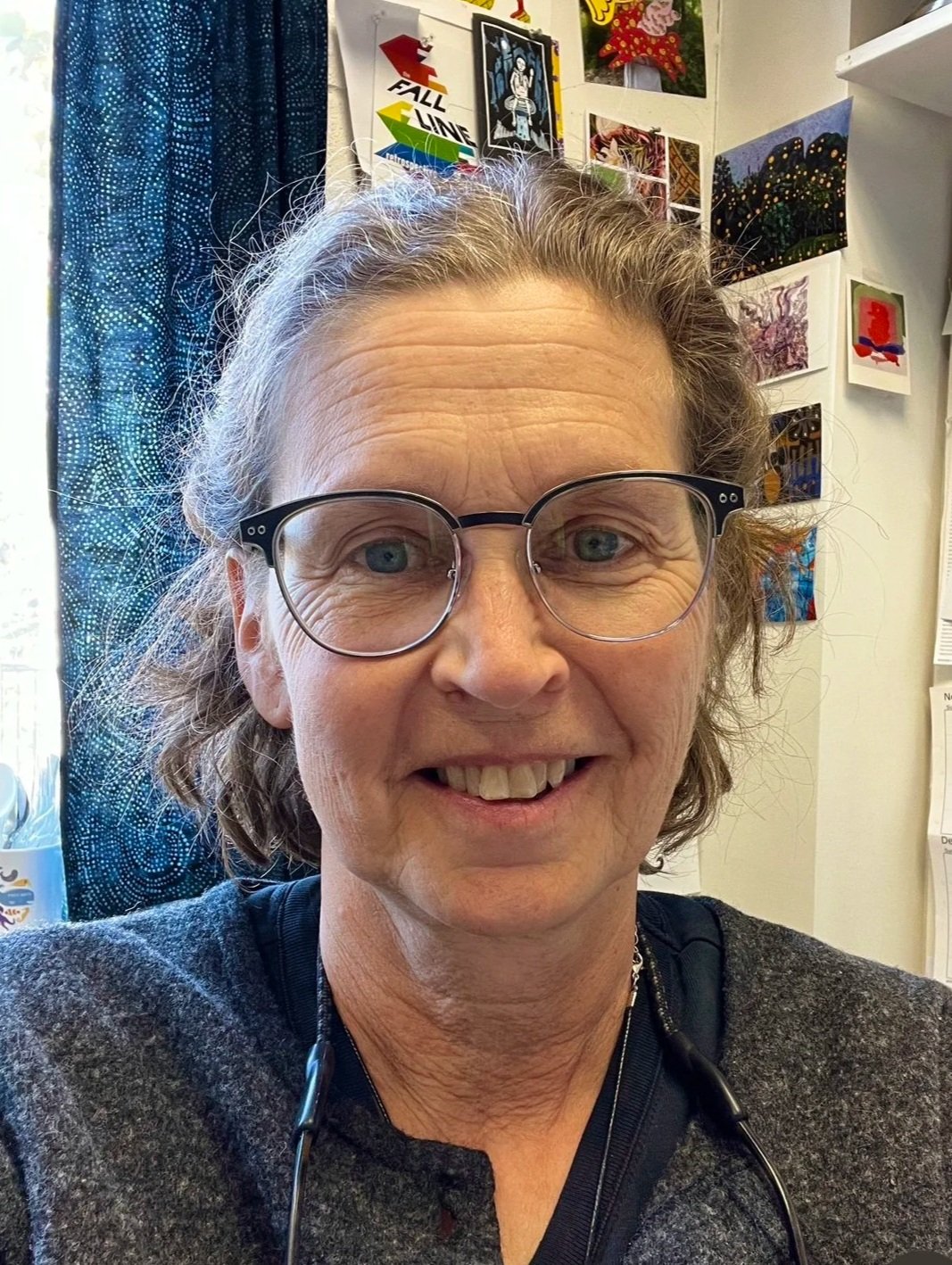 A woman with glasses and gray hair smiling, with colorful wall art and posters in the background.