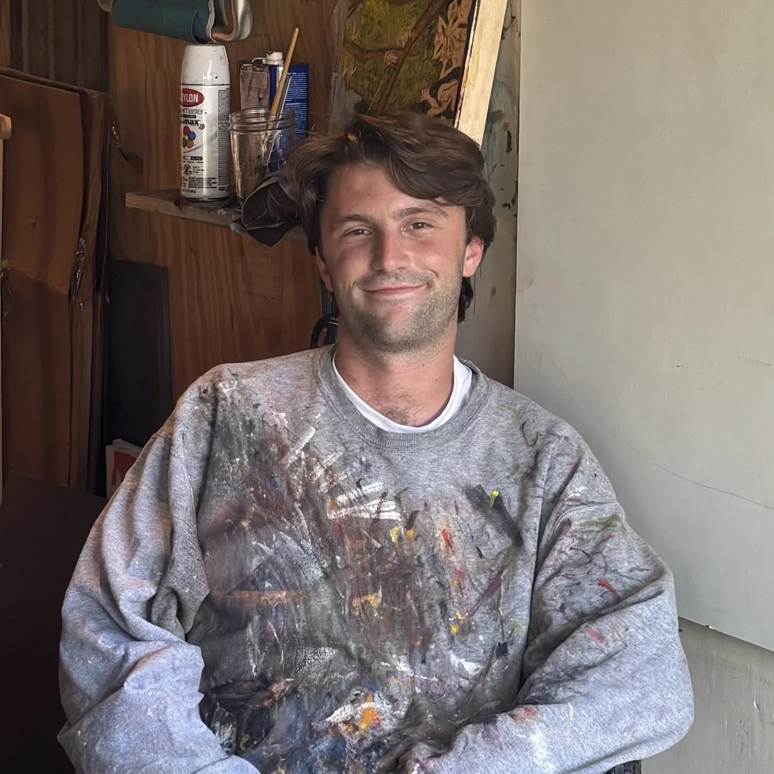 A man with brown hair and a beard sitting indoors, wearing a paint-splattered gray sweatshirt, smiling at the camera.