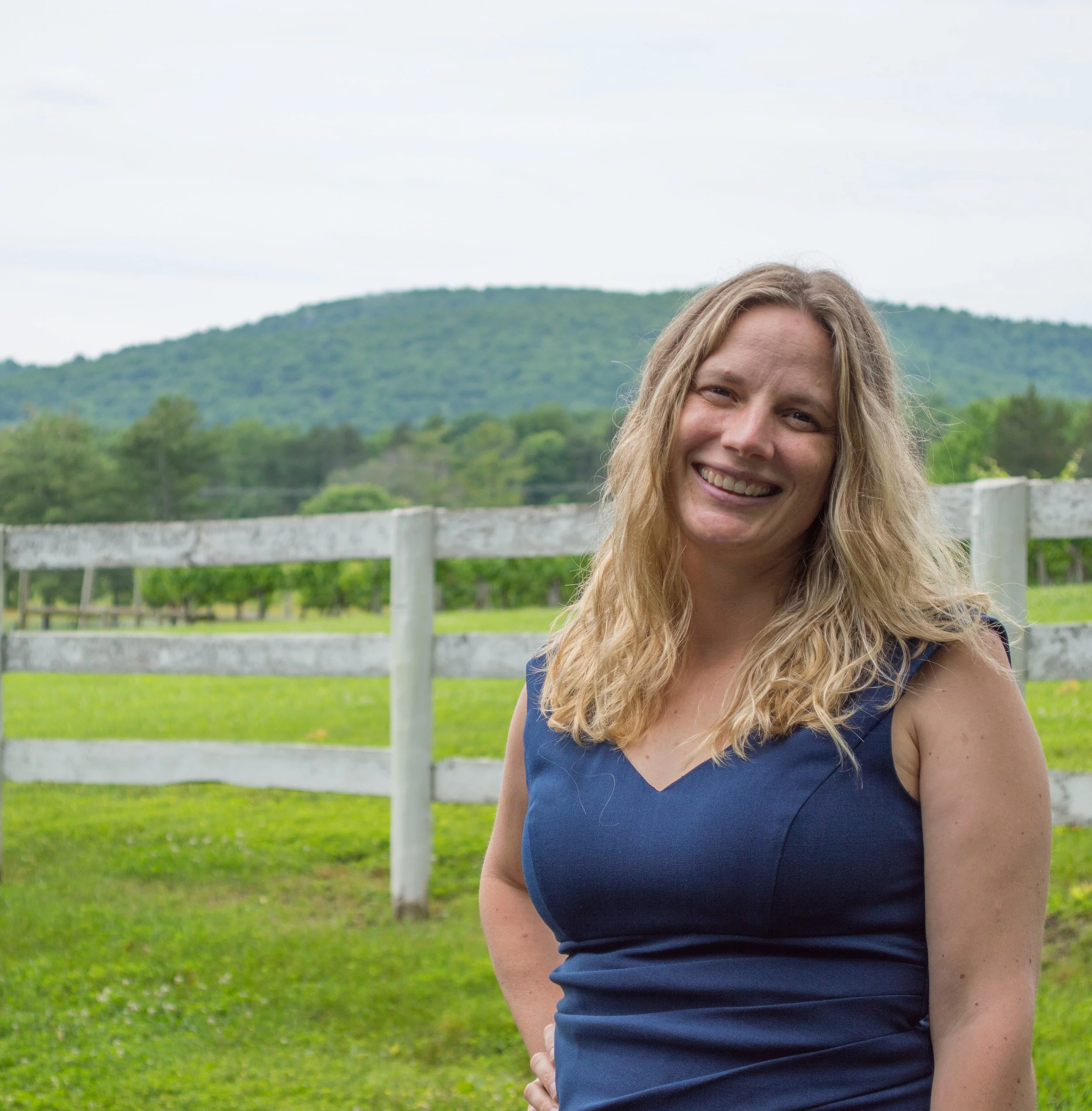 A smiling woman with long blonde hair wearing a dark blue sleeveless top standing outdoors against a backdrop of green trees, a white fence, and distant hills under a cloudy sky.