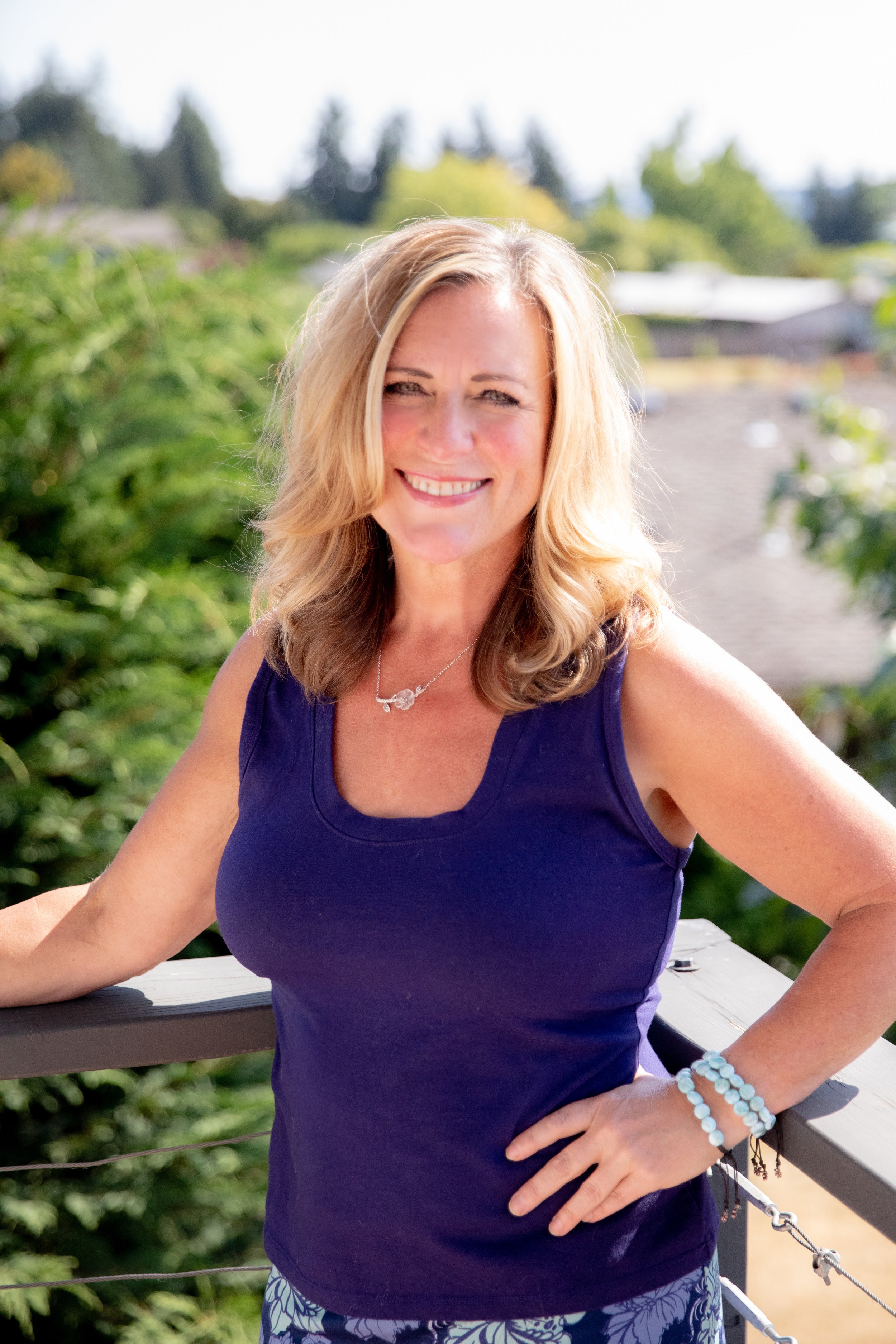 A woman with blonde hair smiling outdoors, wearing a navy sleeveless top, standing on a wooden balcony with green trees and houses in the background.