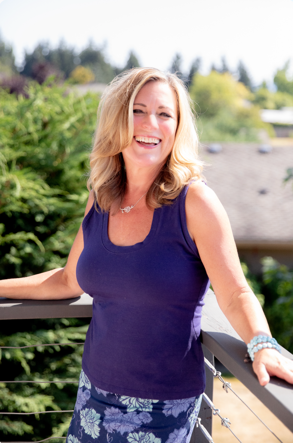A woman with blonde hair wearing a purple sleeveless top and floral skirt, smiling on a balcony with trees and houses in the background.
