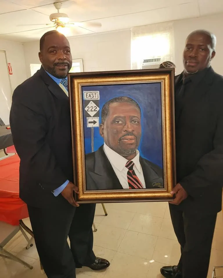 Two men in suits holding a framed portrait of a man in a suit and tie, standing in a room with white walls and a window in the background.