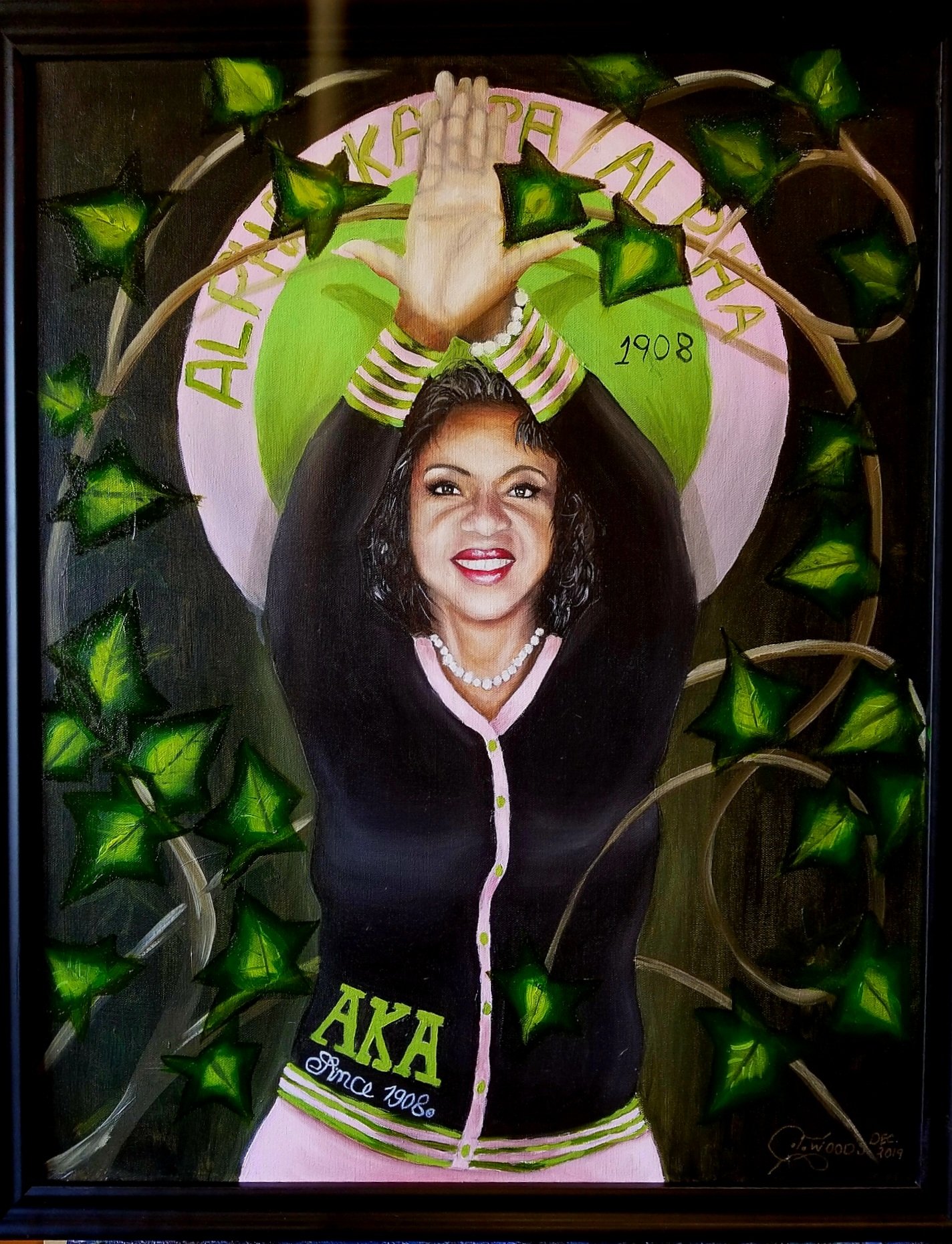 Portrait of a woman with dark curly hair, smiling and raising her hands above her head, surrounded by green ivy leaves, with a sign that reads 'ALPHA KAPPA ALPHA' and 'since 1908'.