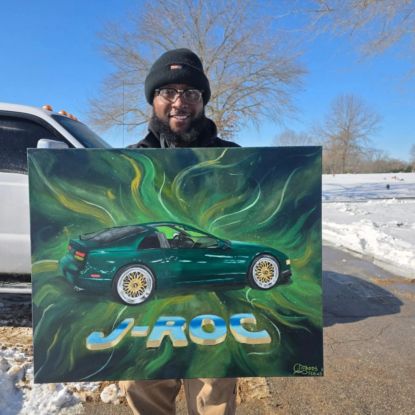 A smiling man wearing glasses and a black beanie stands outdoors in winter, holding a colorful painting of a green sports car with the letters JROC below it, against a backdrop of snow and leafless trees.