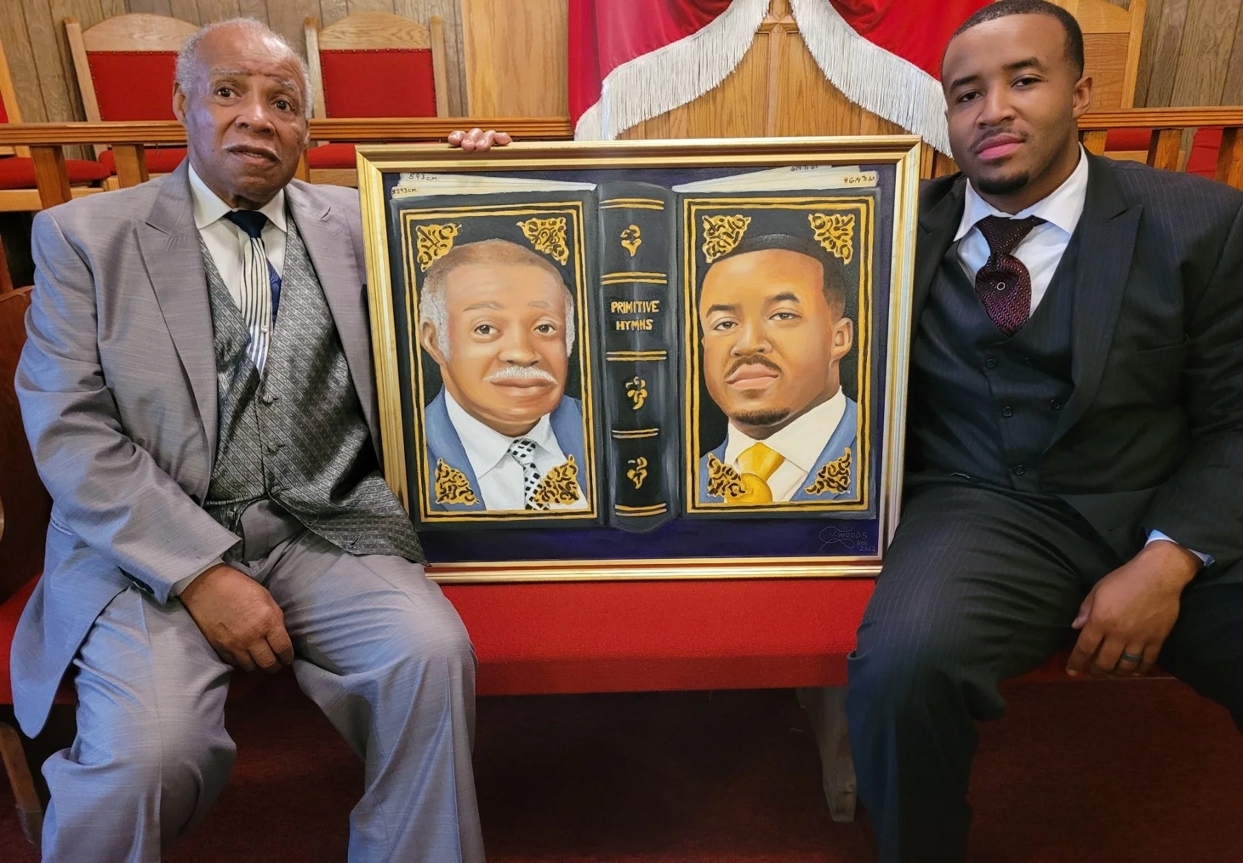 Two men in suits sitting on a red church pew, holding a framed painting of two other men, with a red and white church decoration behind them.