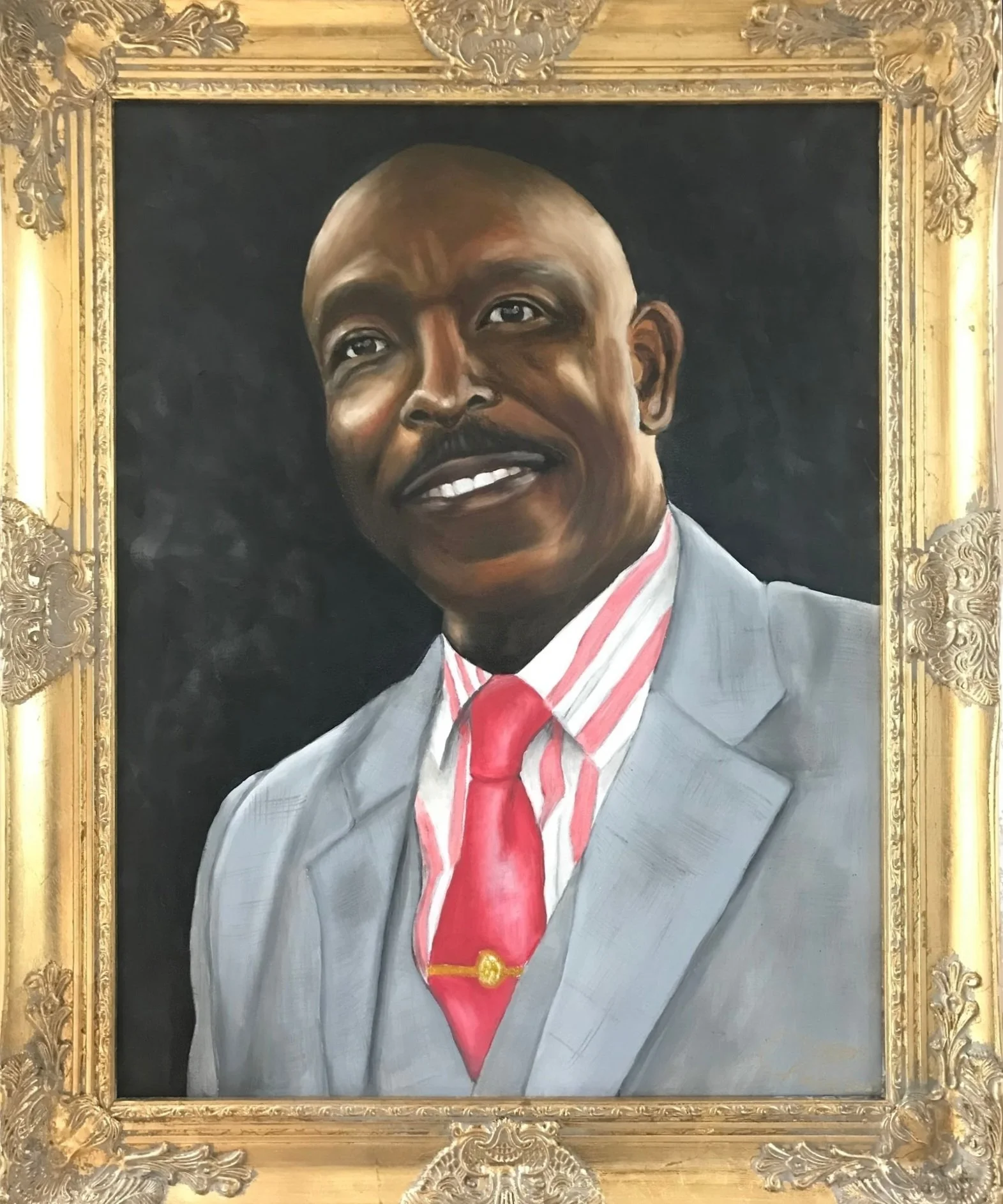 Portrait of a man with a mustache and a bald head, wearing a light gray suit, a red and white striped shirt, and a red tie, framed in an ornate gold frame.