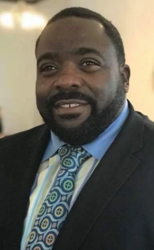 A man with dark skin, a beard, and short hair, wearing a suit and tie, smiling indoors.