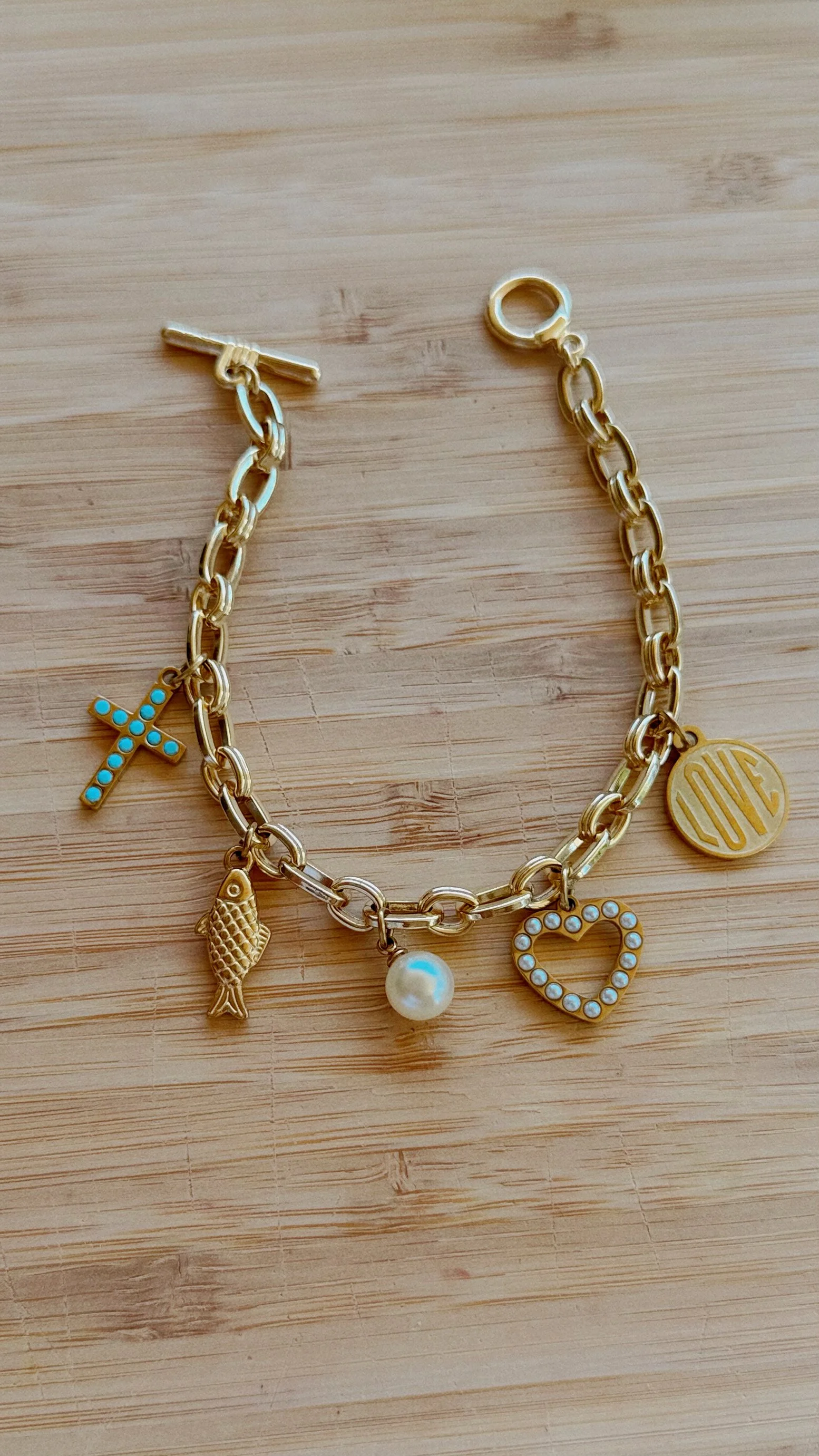 Gold charm bracelet with cross, fish, pearl, heart, and love charms on chain, styled on wood background with greenery