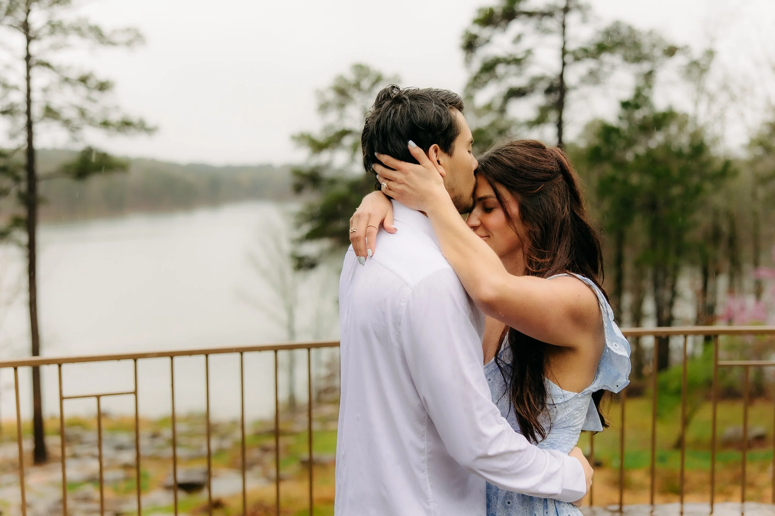 A couple embracing outdoors near a body of water, with trees in the background.