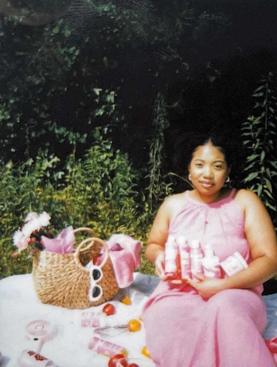 A woman in a pink dress sitting outdoors at a picnic, holding bottles of skincare products, with a woven basket and snacks on a white blanket nearby.