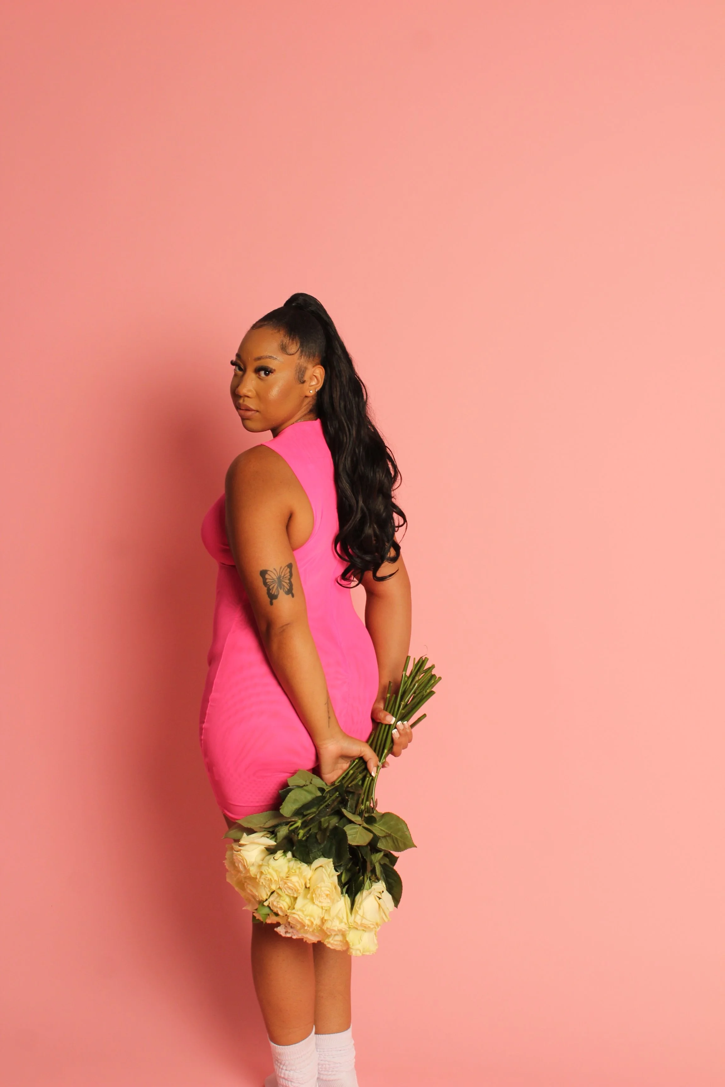 A woman with long black hair in a high ponytail wearing a pink sleeveless dress, holding a bouquet of white roses, standing against a pink background.