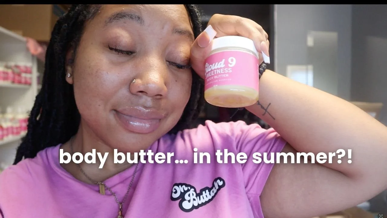 A woman with closed eyes holding a jar of body butter near her face, wearing a pink shirt with a patch that says "Dr. Body Butter," and standing in a room with shelves of products in the background.