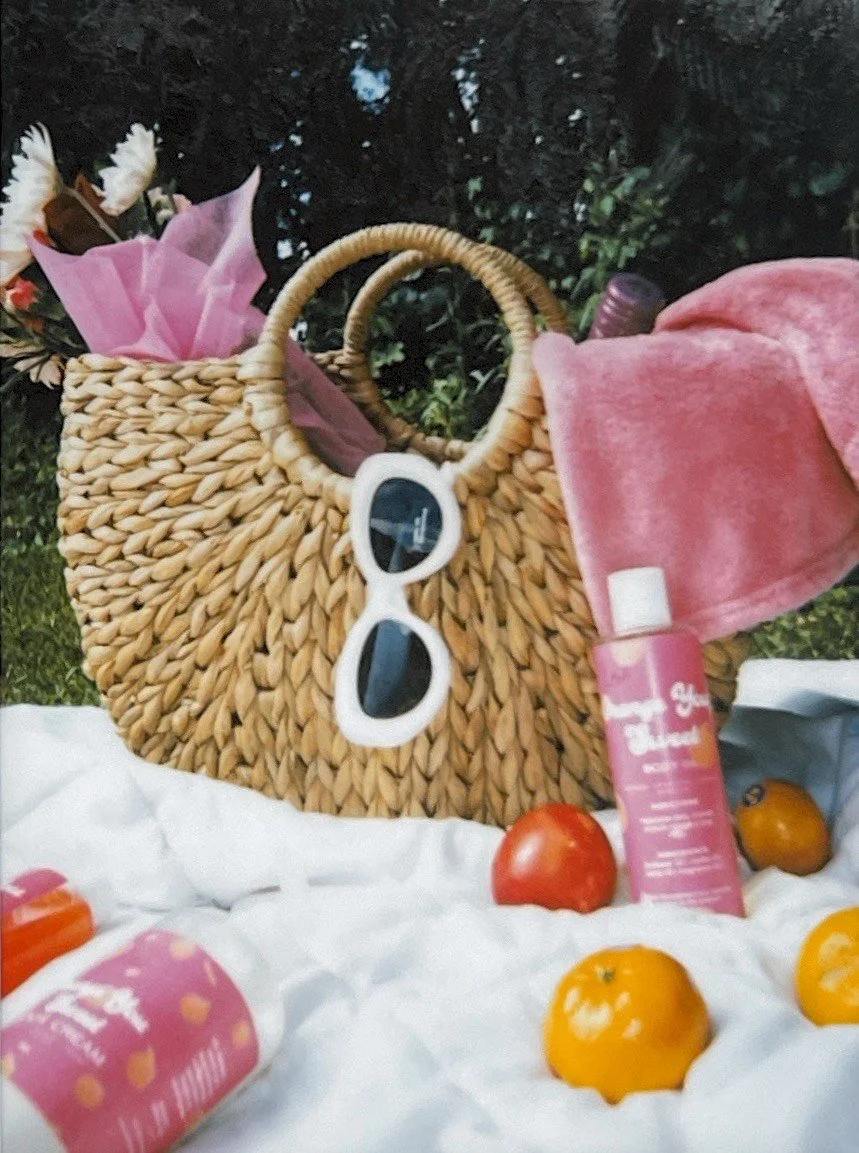 A woven straw handbag with white sunglasses hanging on it, filled with pink and white flowers, sitting on a white cloth with tomatoes and skincare products nearby, outdoors with greenery in the background.