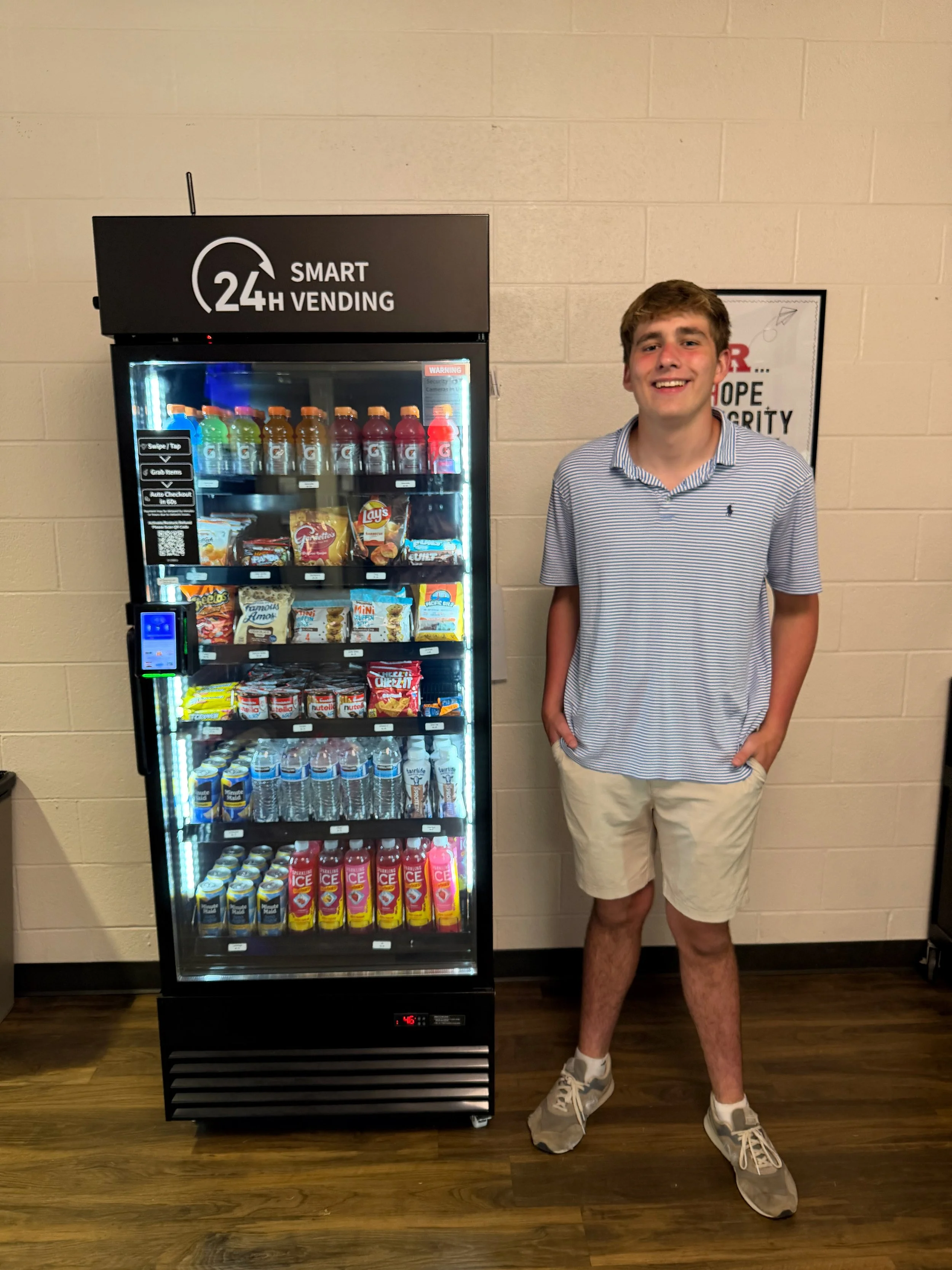 A young man in a striped polo shirt and beige shorts standing next to a vending machine with snacks and drinks, smiling.