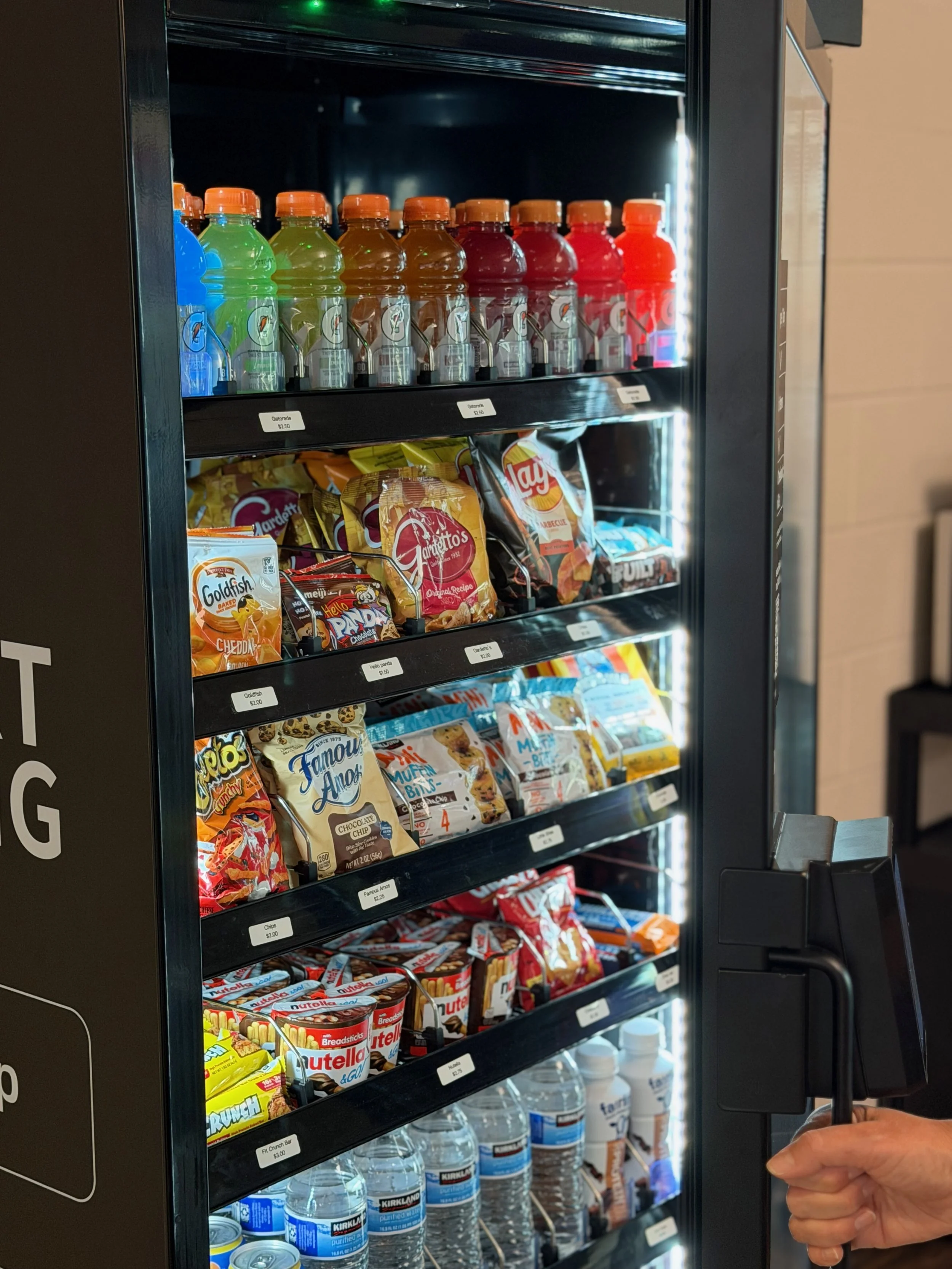 Vending machine stocked with colorful bottled drinks, snack bags including chips and cookies, Nutella, and bottled water.