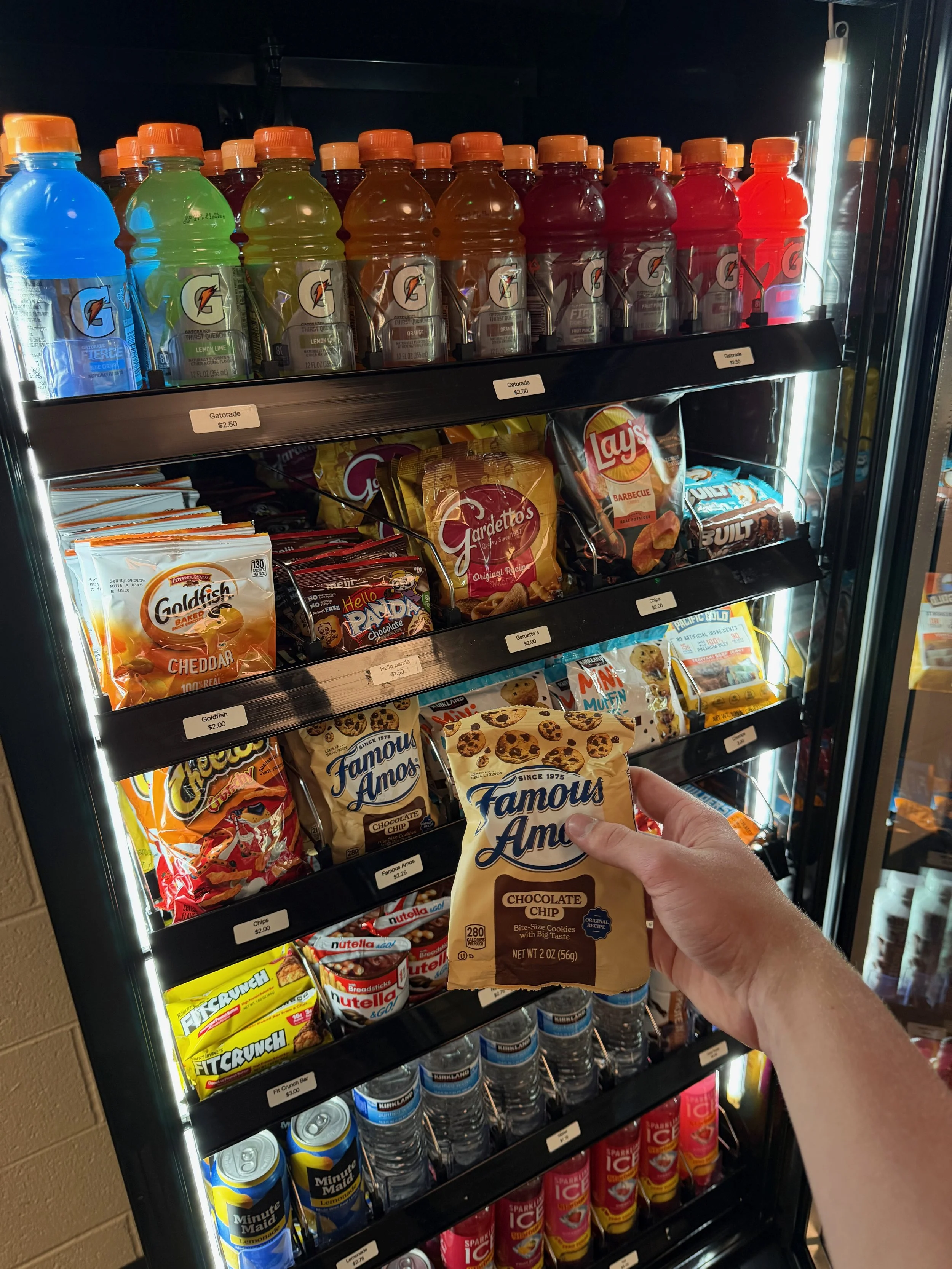Hand holding a bag of Famous Amos chocolate chip cookies in front of a vending machine with snacks and drinks.