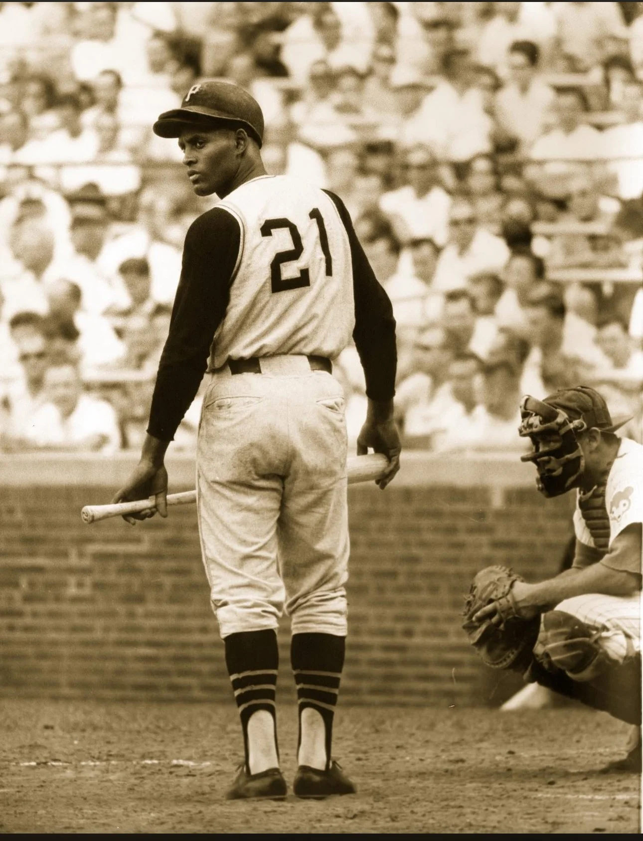 Historical black and white photo of a baseball player holding a bat, wearing a uniform with the number 21, standing on the field. Another player, dressed as a catcher, is crouched nearby, with a crowd in the background.