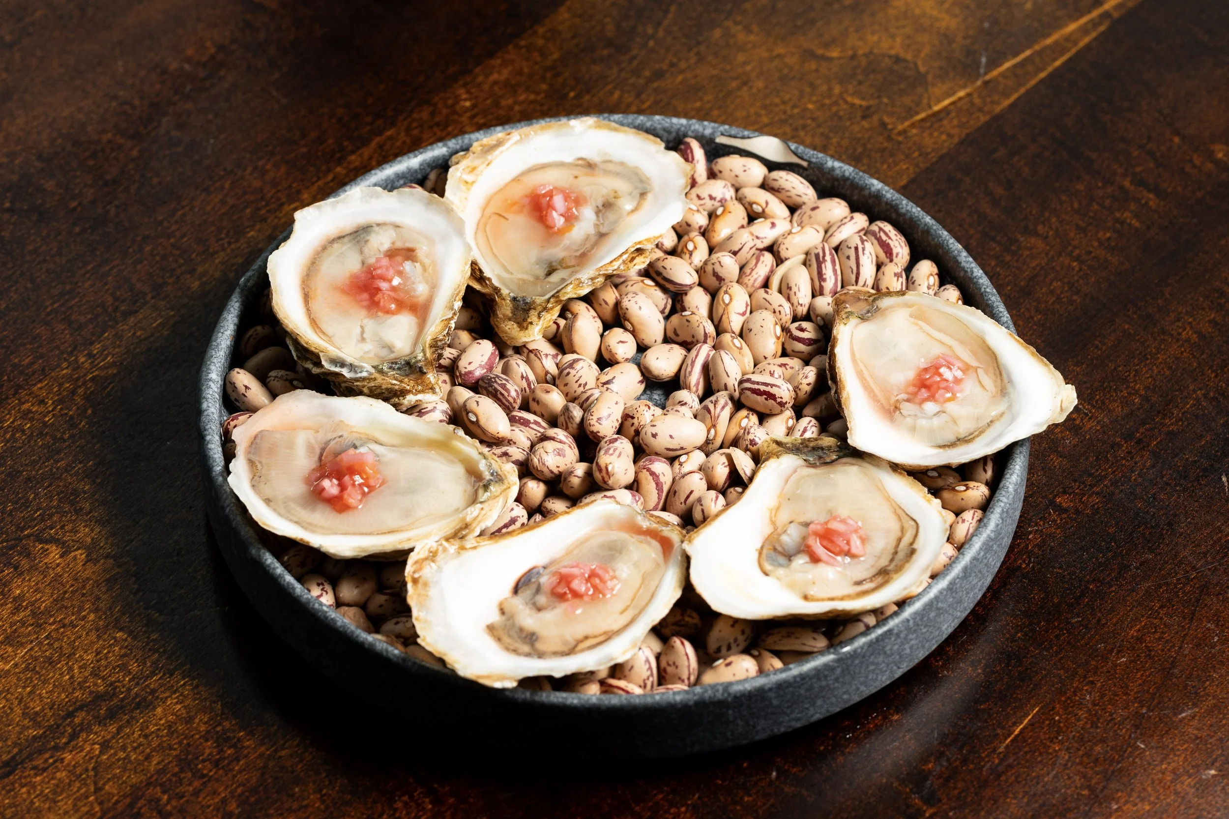 A black bowl containing cooked oysters with red roe, placed on a wooden table.